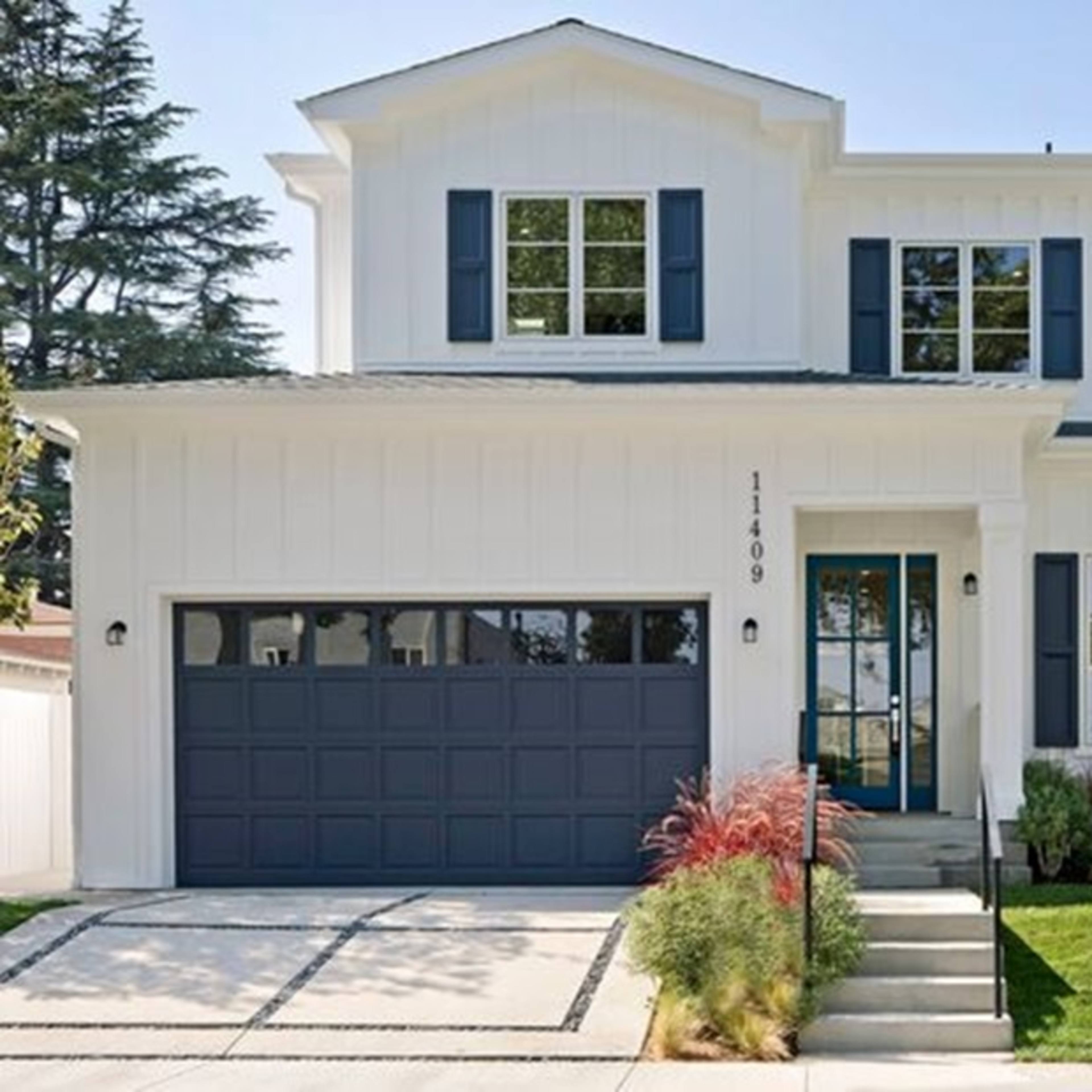 A two-story white house with a dark garage door and blue shutters, surrounded by neatly maintained landscaping.