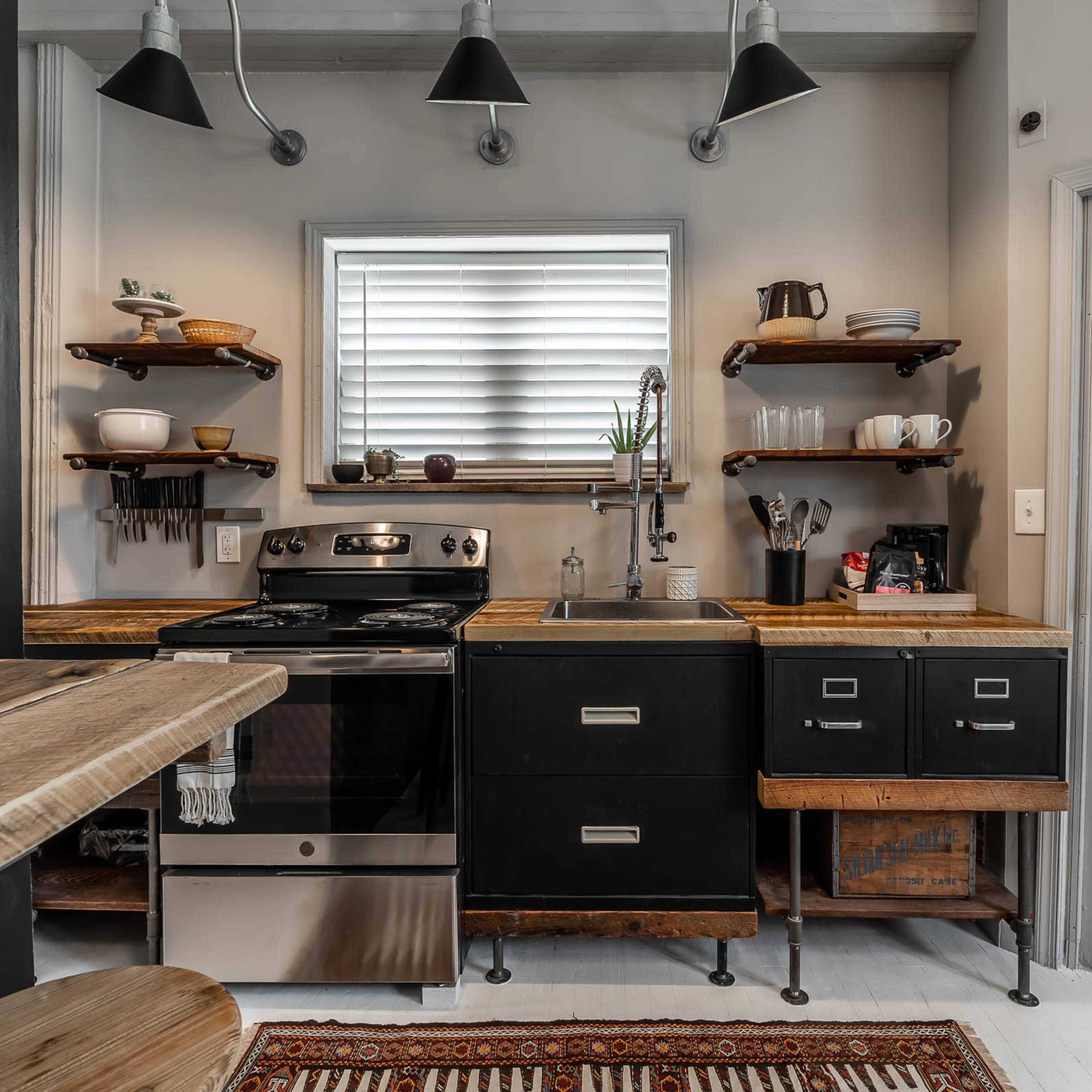 A compact kitchen with black cabinets, wooden shelves, a stainless steel stove, and a farmhouse-style sink.
