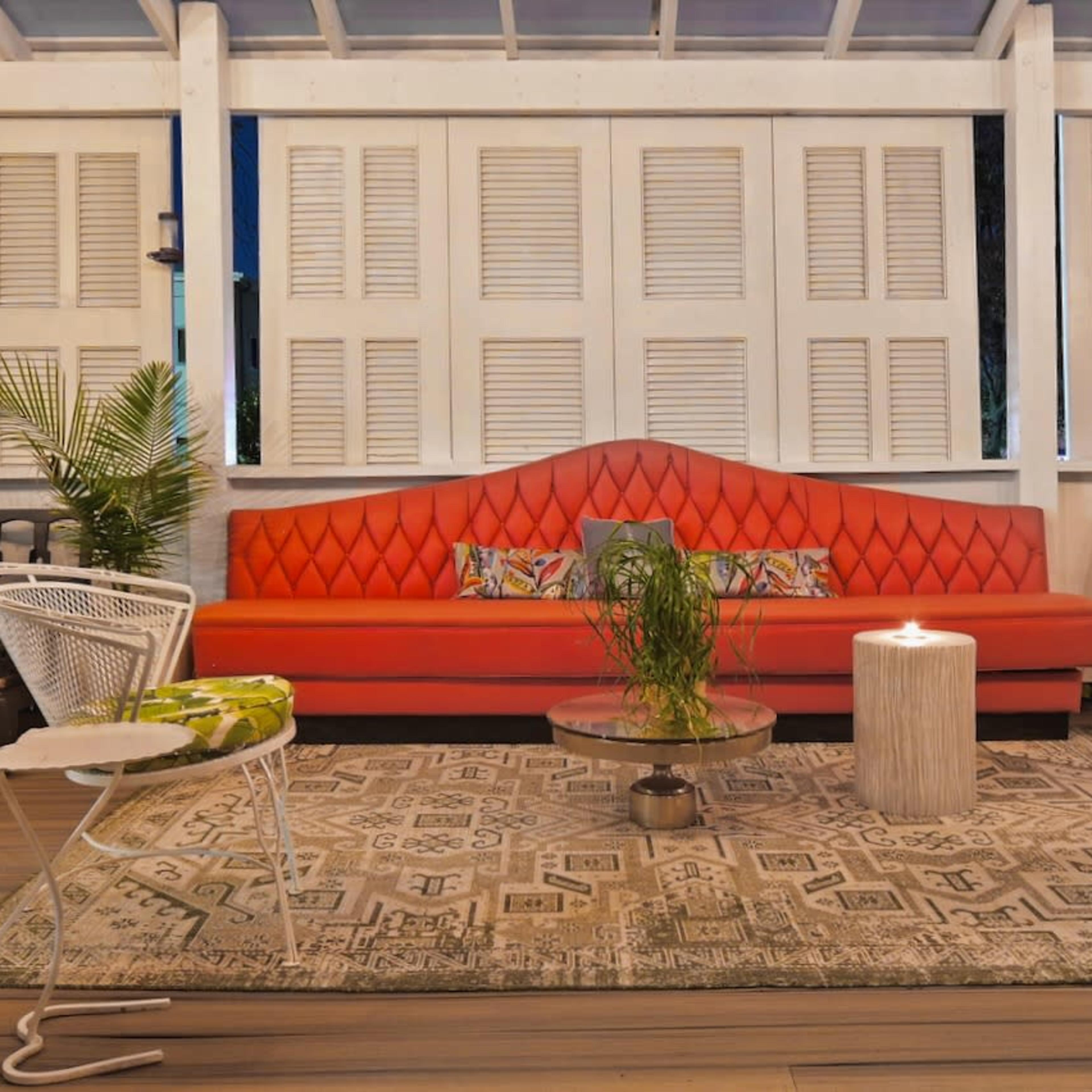 A vibrant red sofa is positioned in a cozy seating area with white wooden shutters, surrounded by decorative plants and a modern coffee table on a patterned rug.