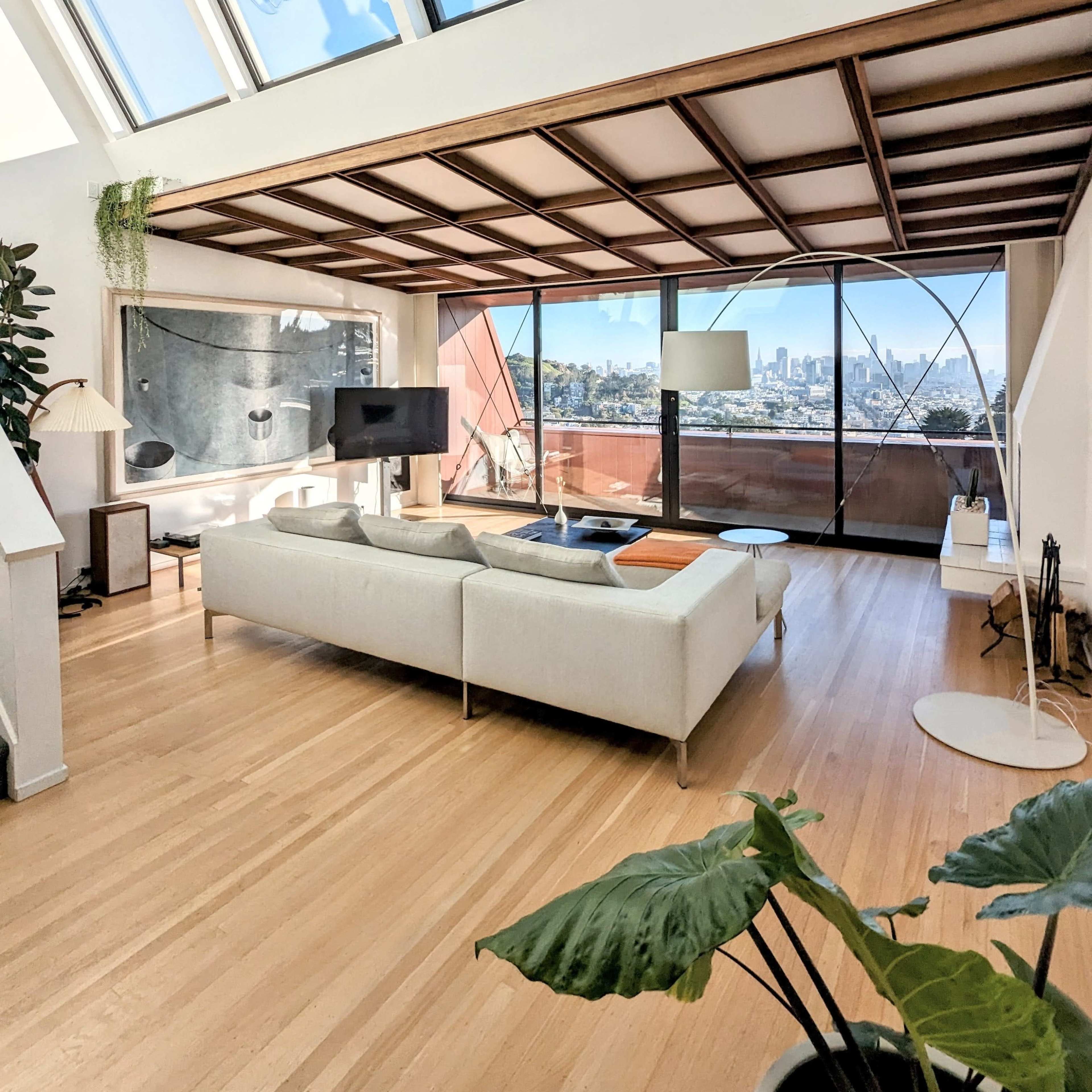 A modern living room with large windows and a view of a city skyline, featuring a light-colored sofa and wooden ceiling beams.