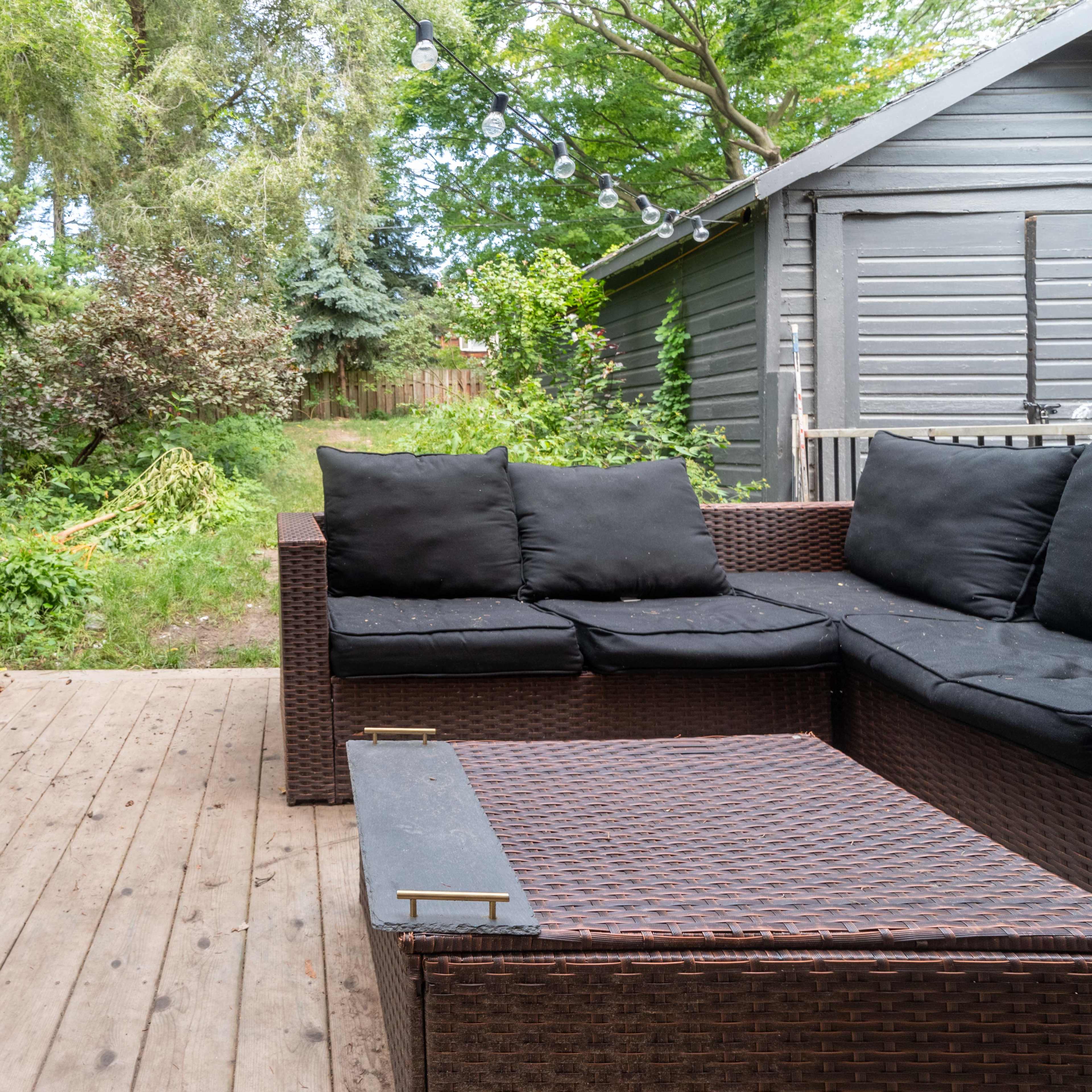 A wooden deck features a dark wicker seating area and a high table with stools, surrounded by greenery and trees.