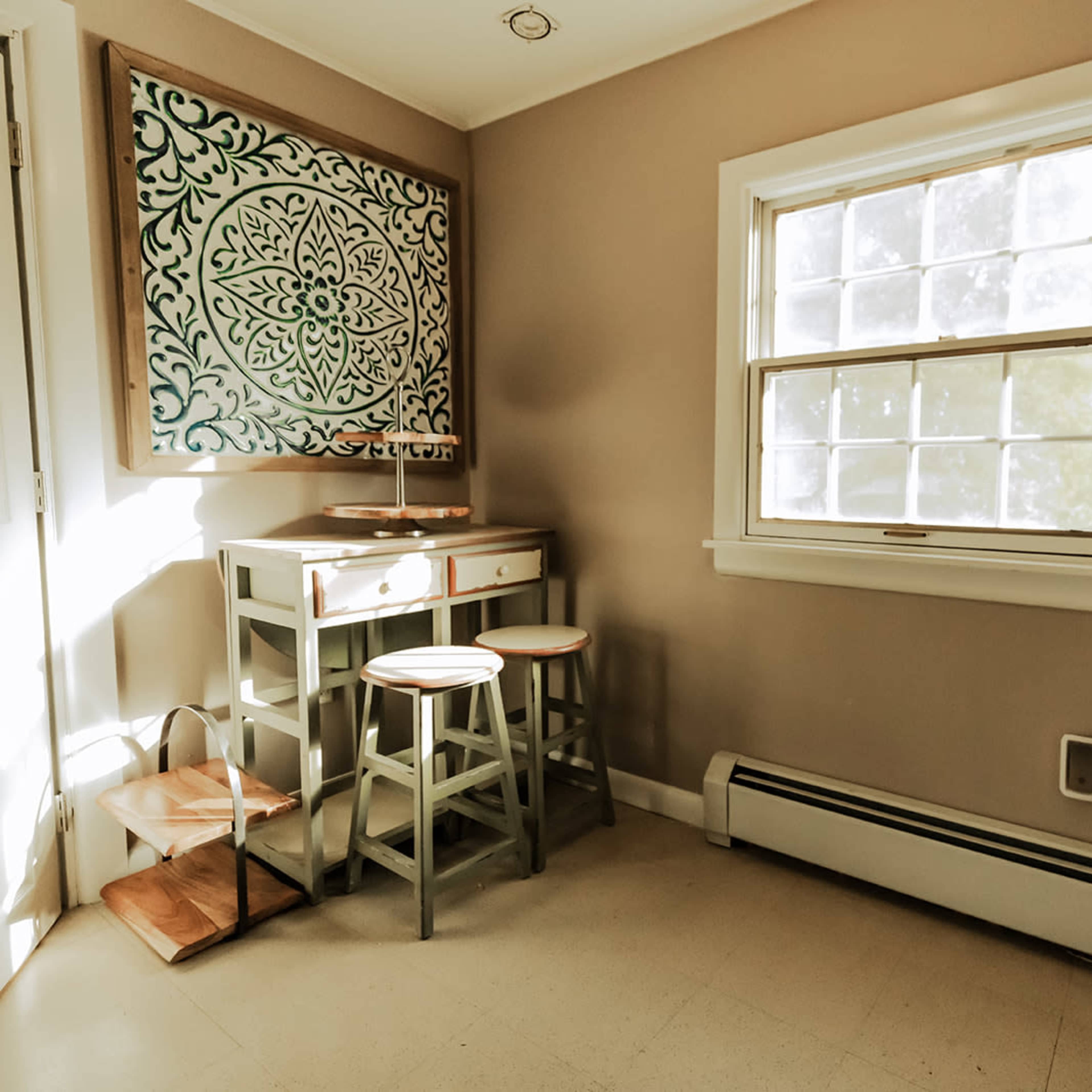 A small room with a window, a wall decoration, a table with two stools, and a radiator.