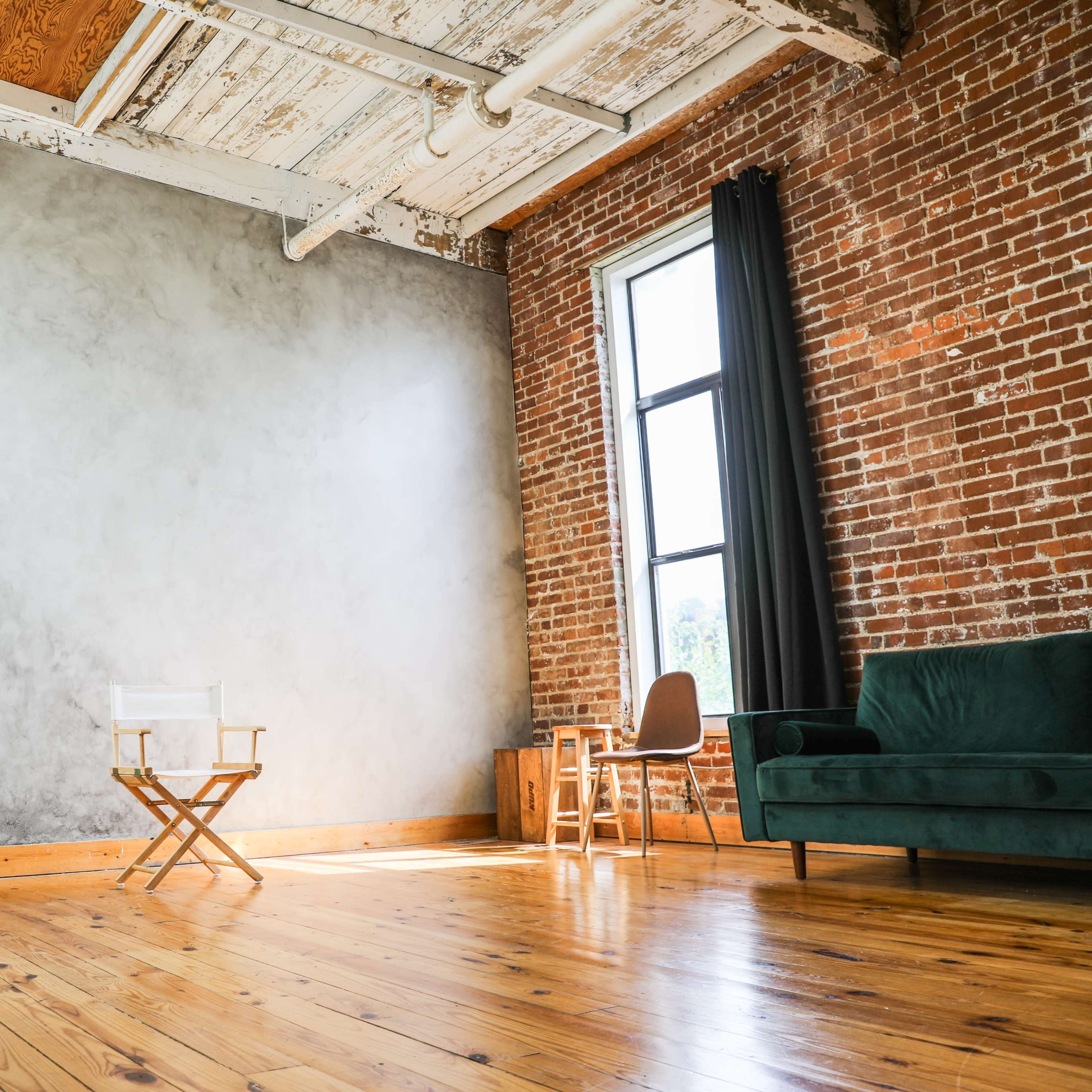 A spacious room with a wooden floor, exposed brick walls, a gray accent wall, a green sofa, a director's chair, and large windows that allow natural light to enter.