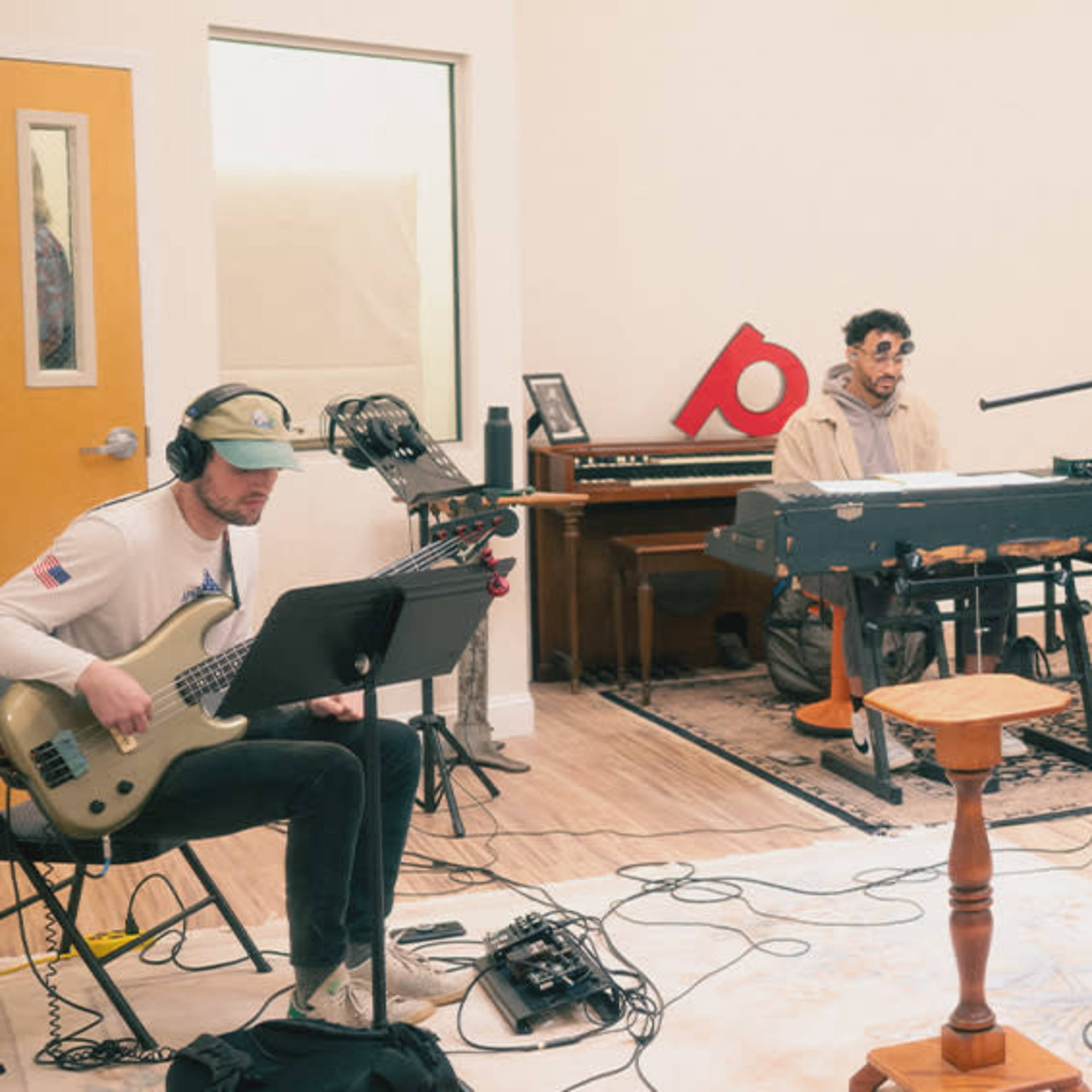 Three musicians are playing instruments in a well-lit recording studio, with a keyboard, a bass guitar, and drums set up around them.