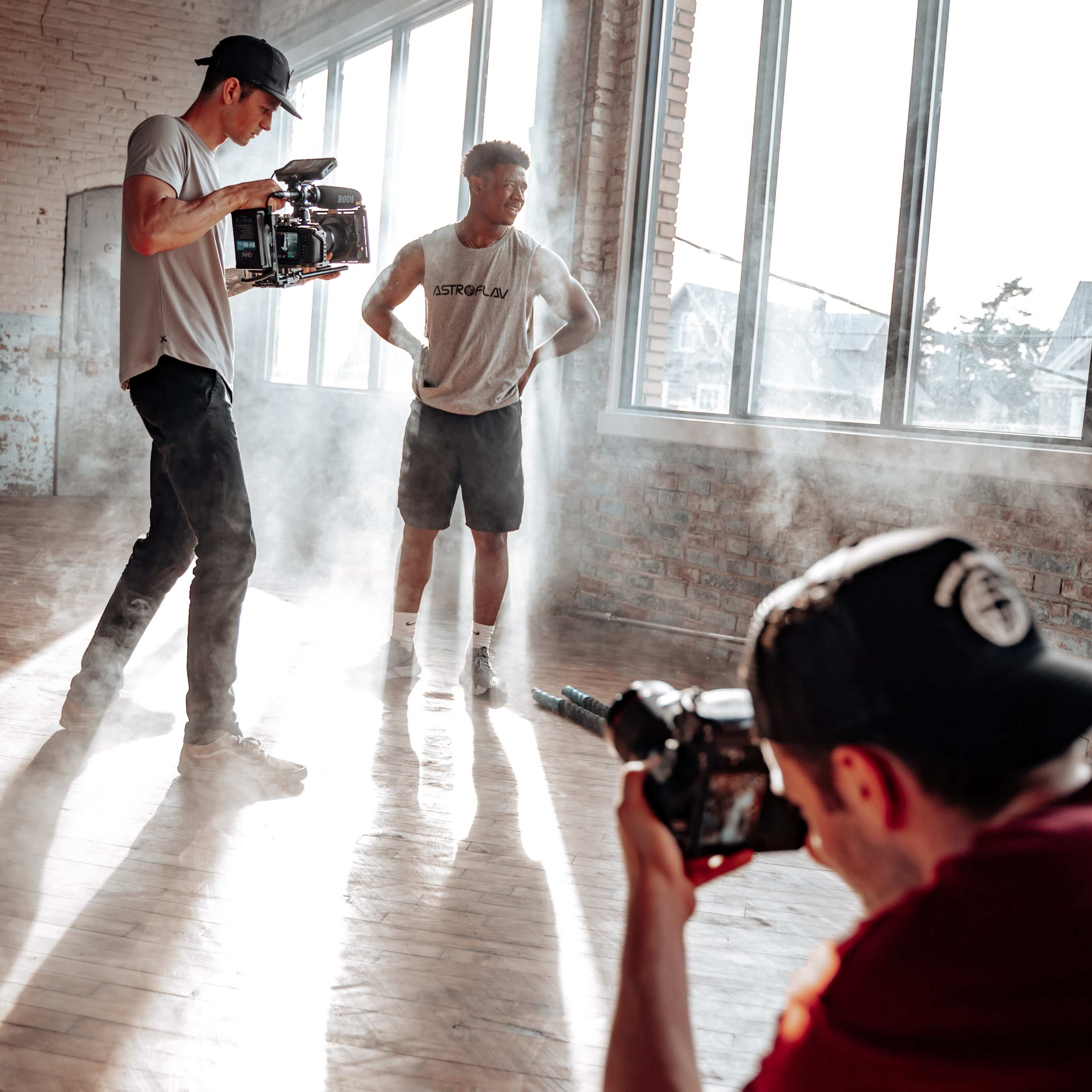 Three individuals are filming a scene in a sunlit, empty room with large windows, while smoke fills the air.
