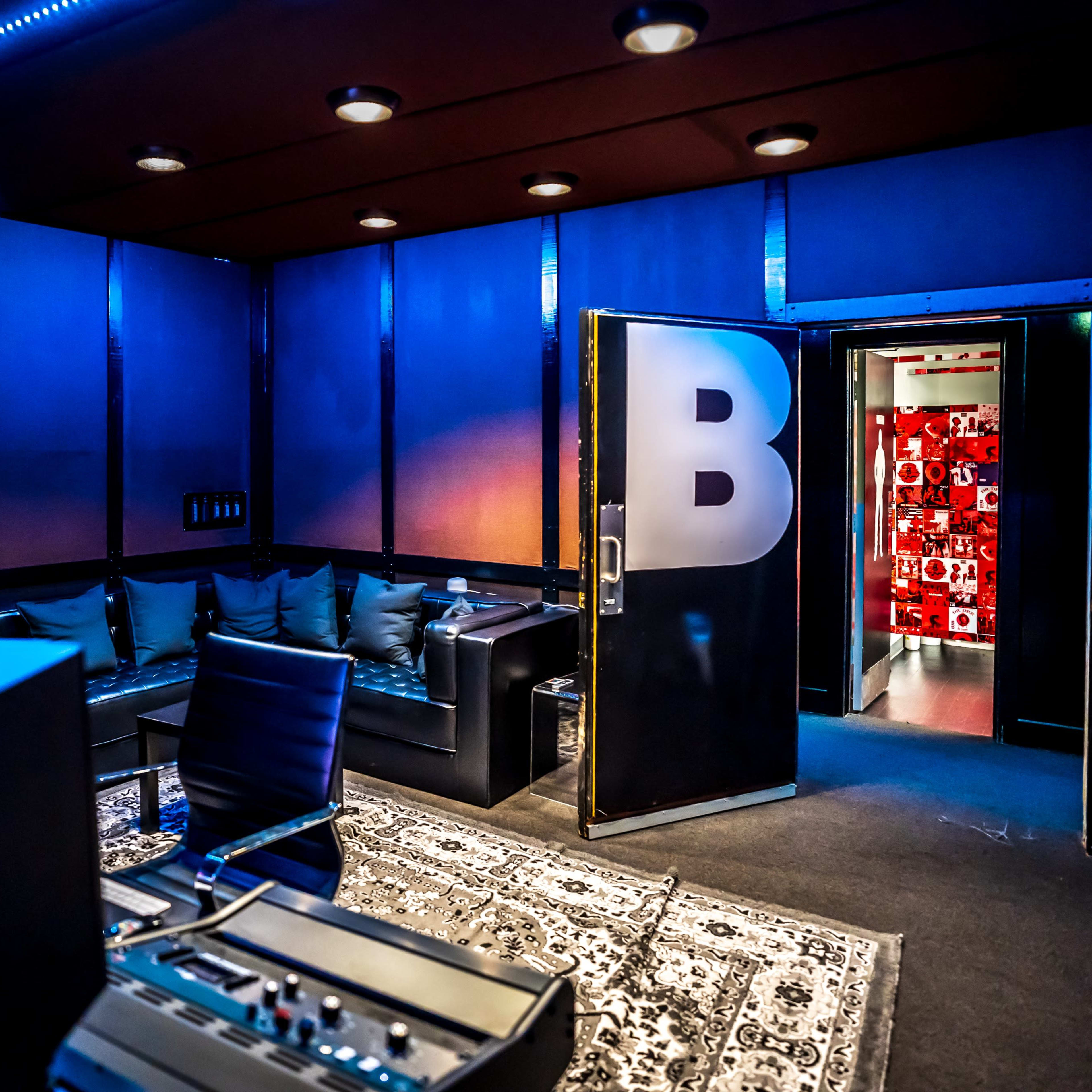 A modern recording studio features a black leather couch and a door marked with a large "B" opening to a vibrant red hallway.