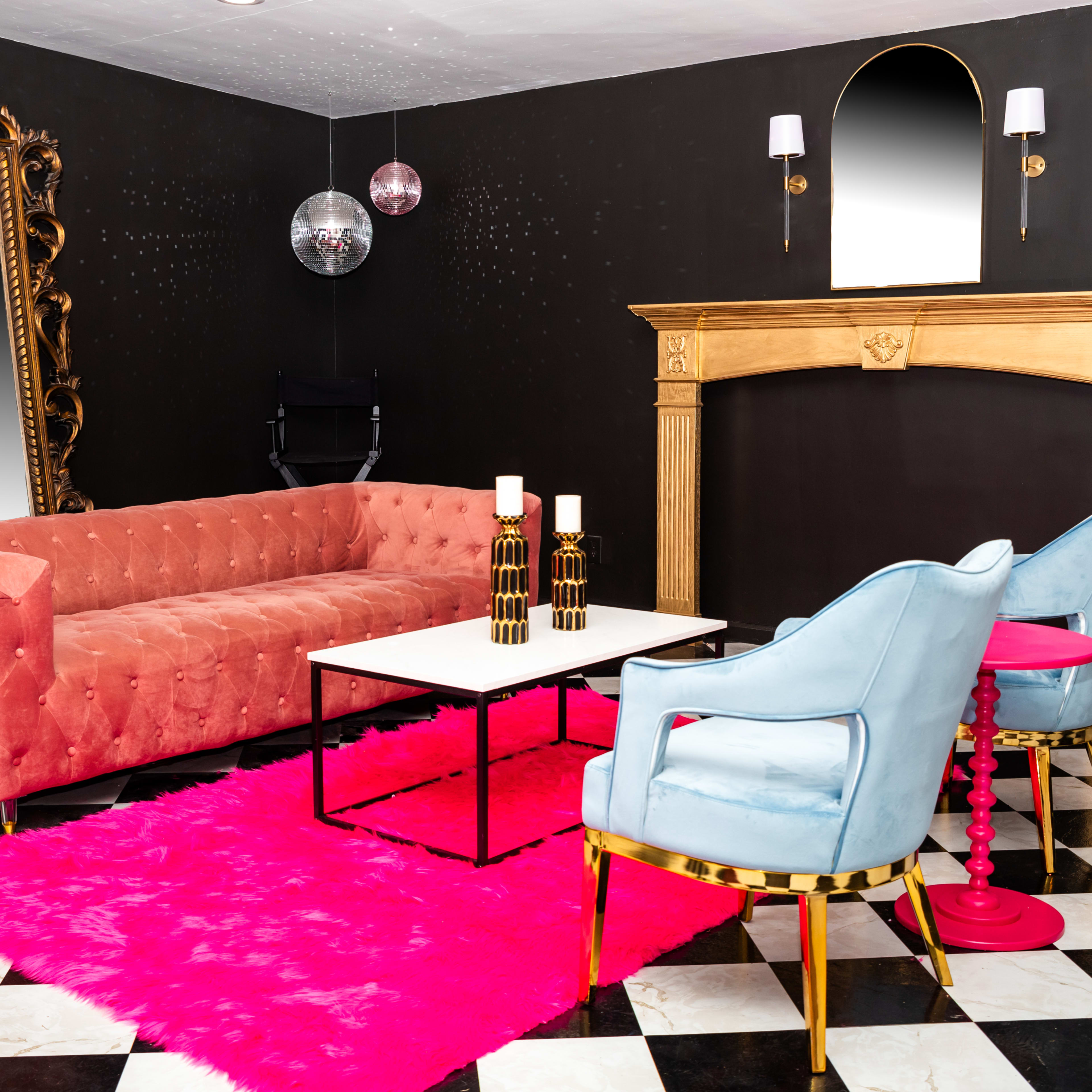 A stylish interior with a pink rug, a tufted sofa, two blue chairs, a marble-topped coffee table, a large ornate mirror, and a golden fireplace against a black wall.