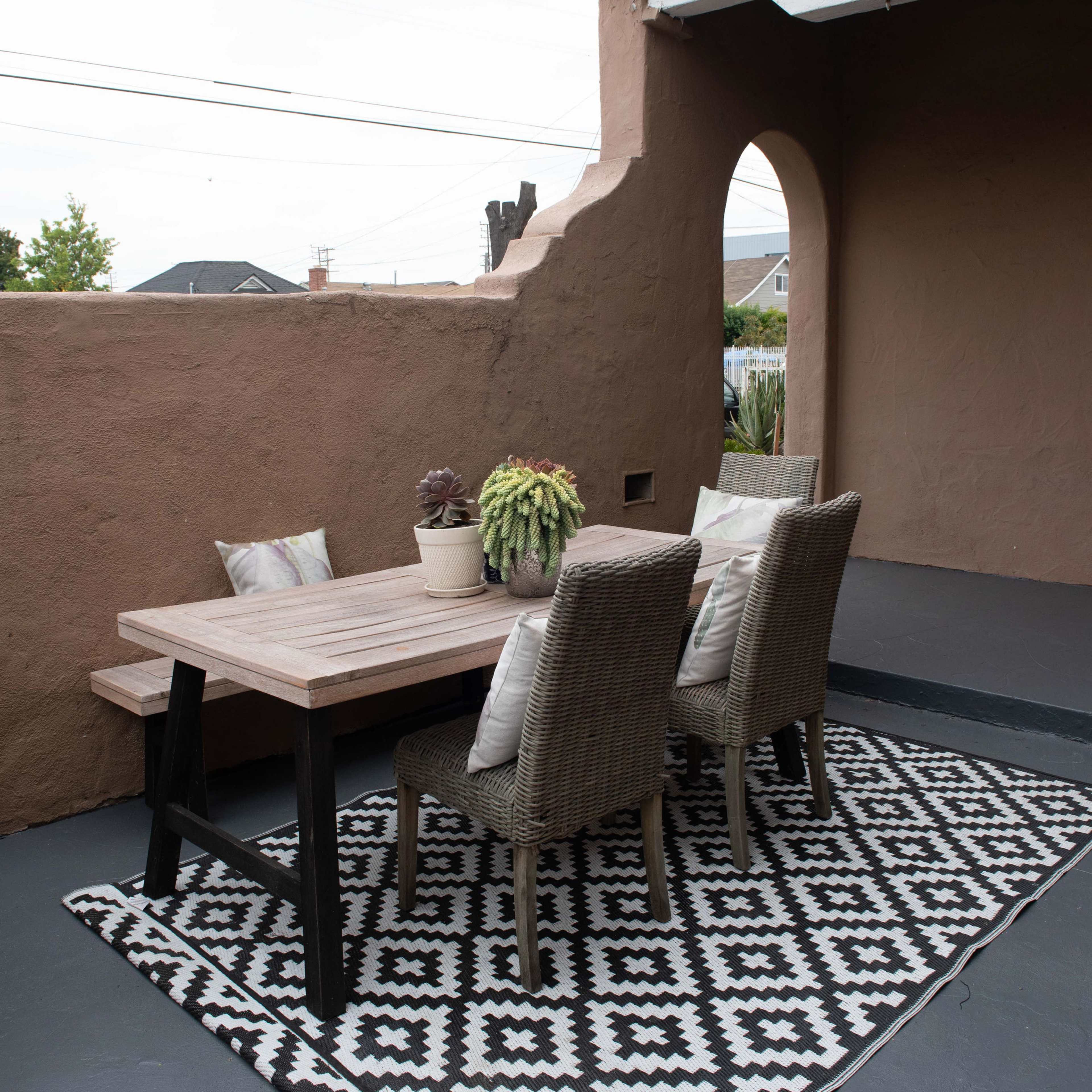 A wooden dining table with two chairs is positioned on a patio covered with a patterned rug, alongside a wall with an archway.