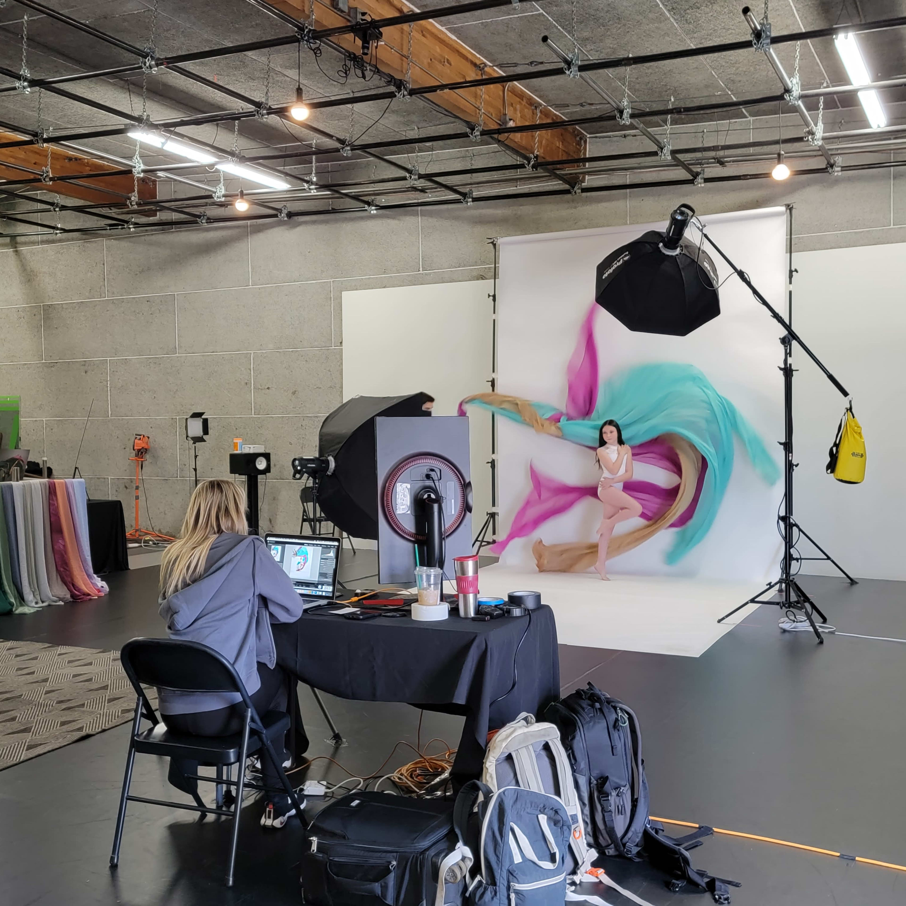 A photographer sets up a photo shoot with a model against a colorful backdrop in a studio with various fabric drapes and lighting equipment.