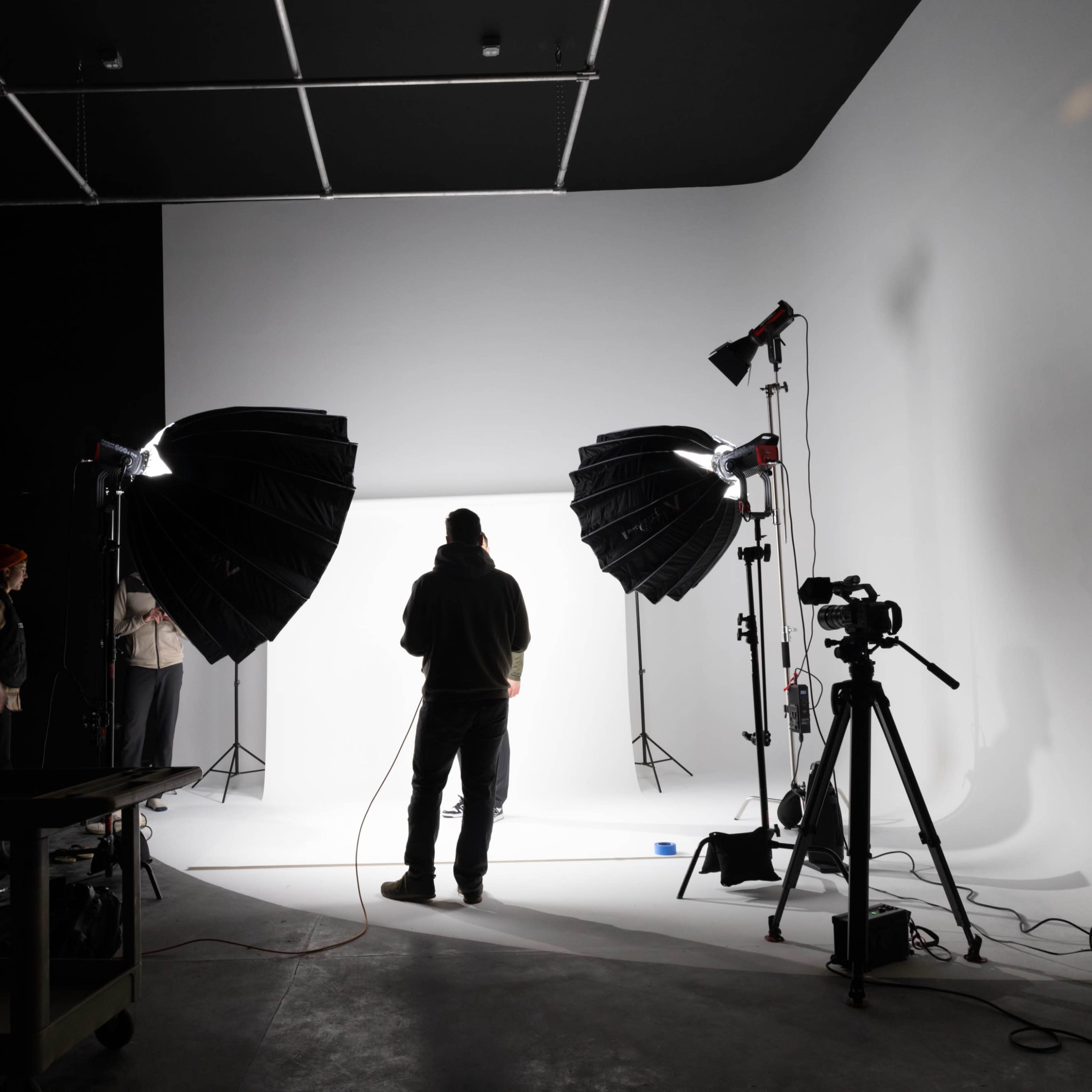 A person stands in front of a backdrop in a photography studio, with softbox lights set up on either side and a camera on a tripod nearby.