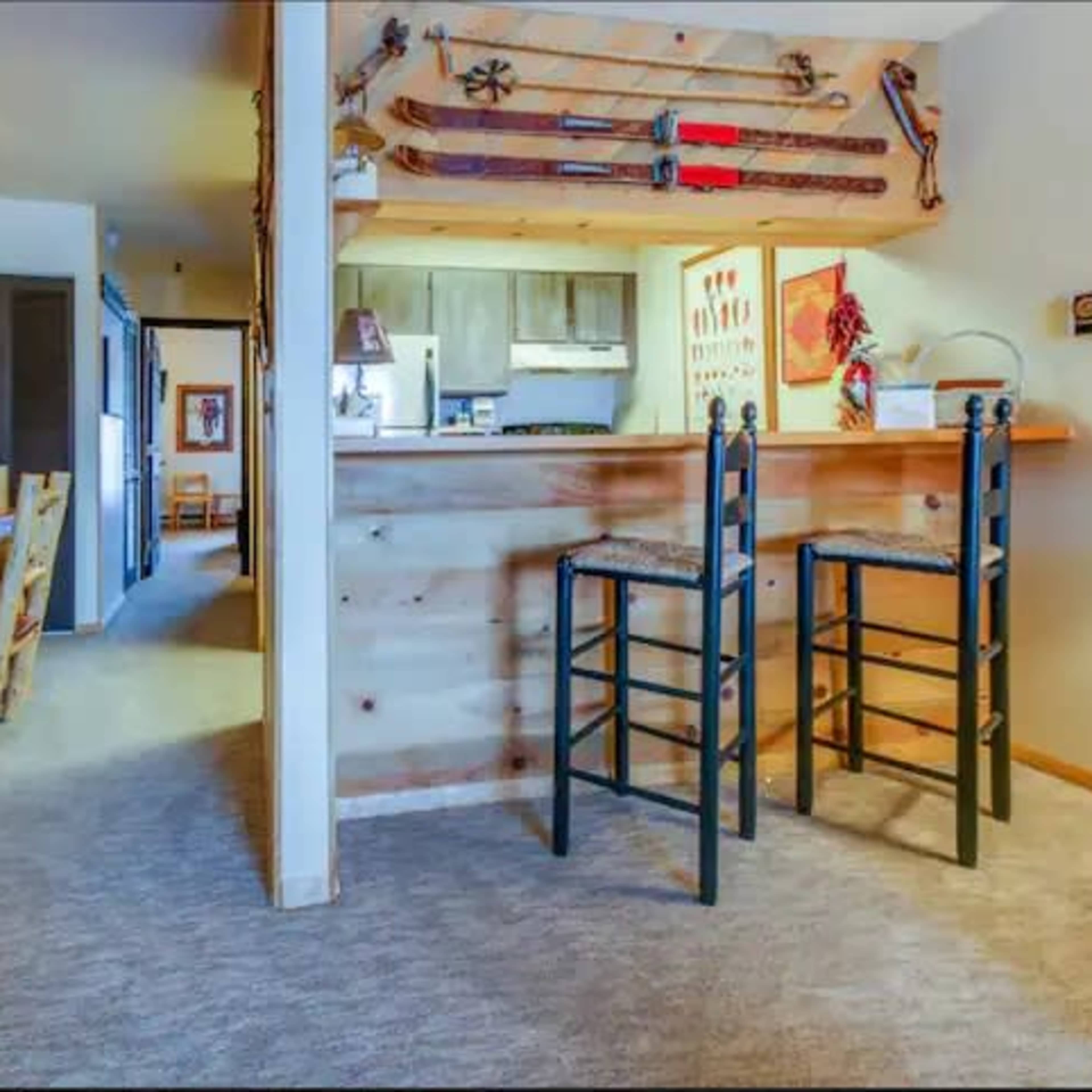 A cozy interior space with wooden walls, a small kitchen area, a bar with two black stools, and rustic decor including skis and snowshoes on display.