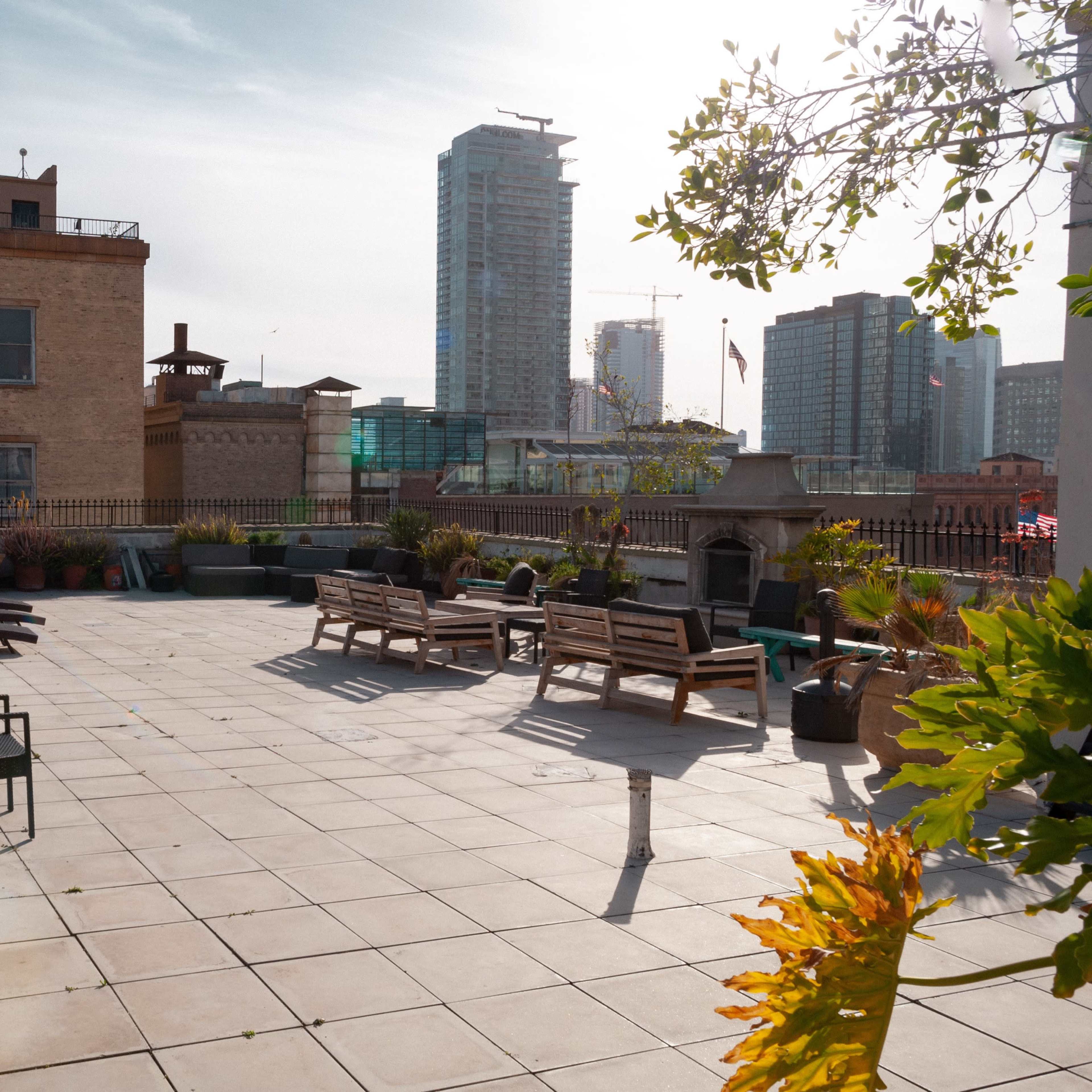 A spacious rooftop terrace with benches and planters, surrounded by city buildings and a clear sky.