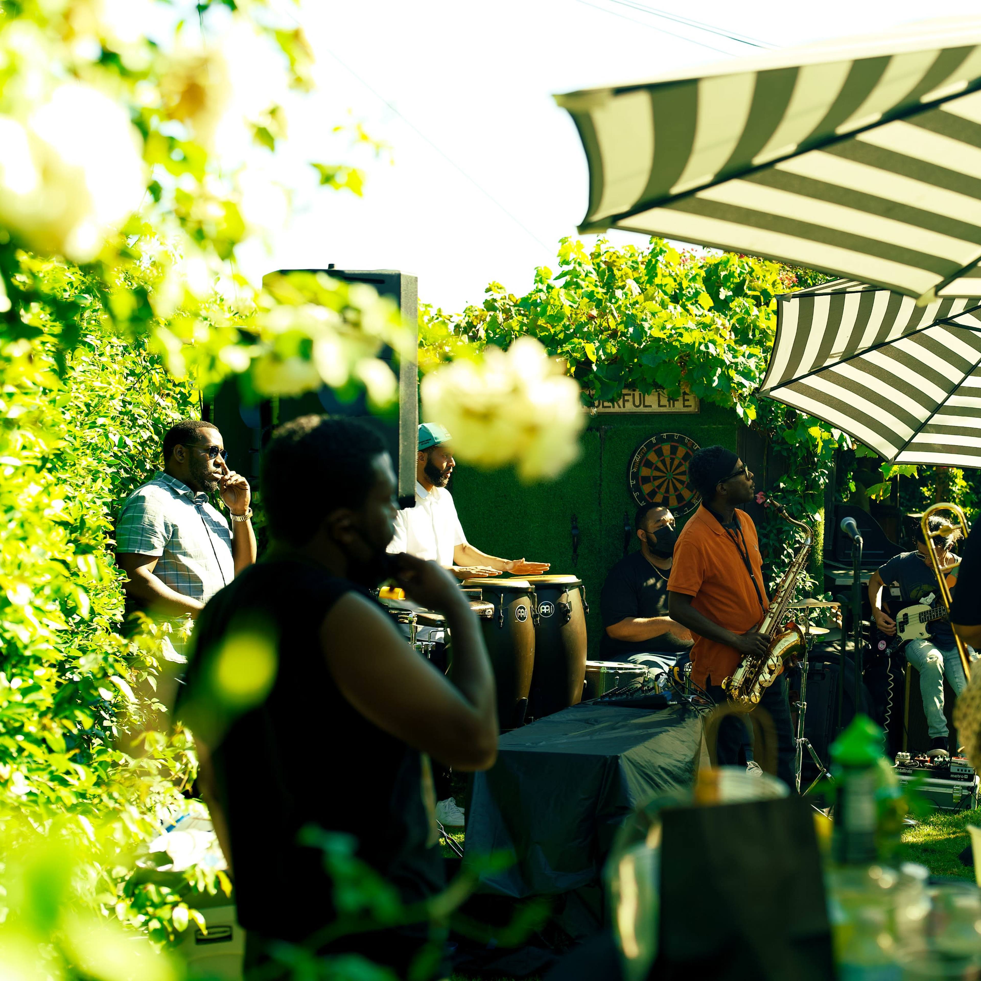A live band performs in a garden while an audience listens under striped umbrellas.