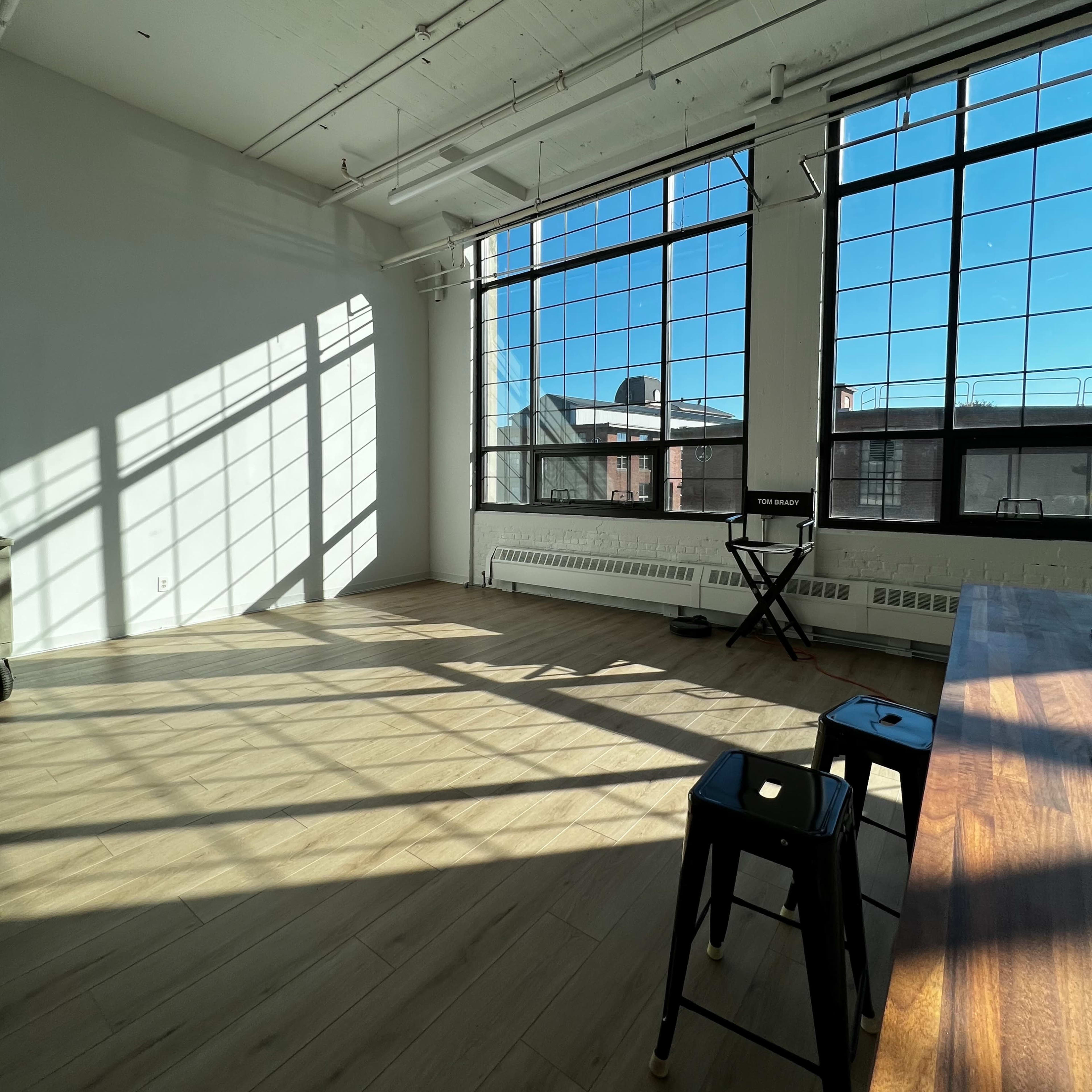 A spacious room with large windows, casting shadows on the wooden floor, and featuring a table and two black stools.