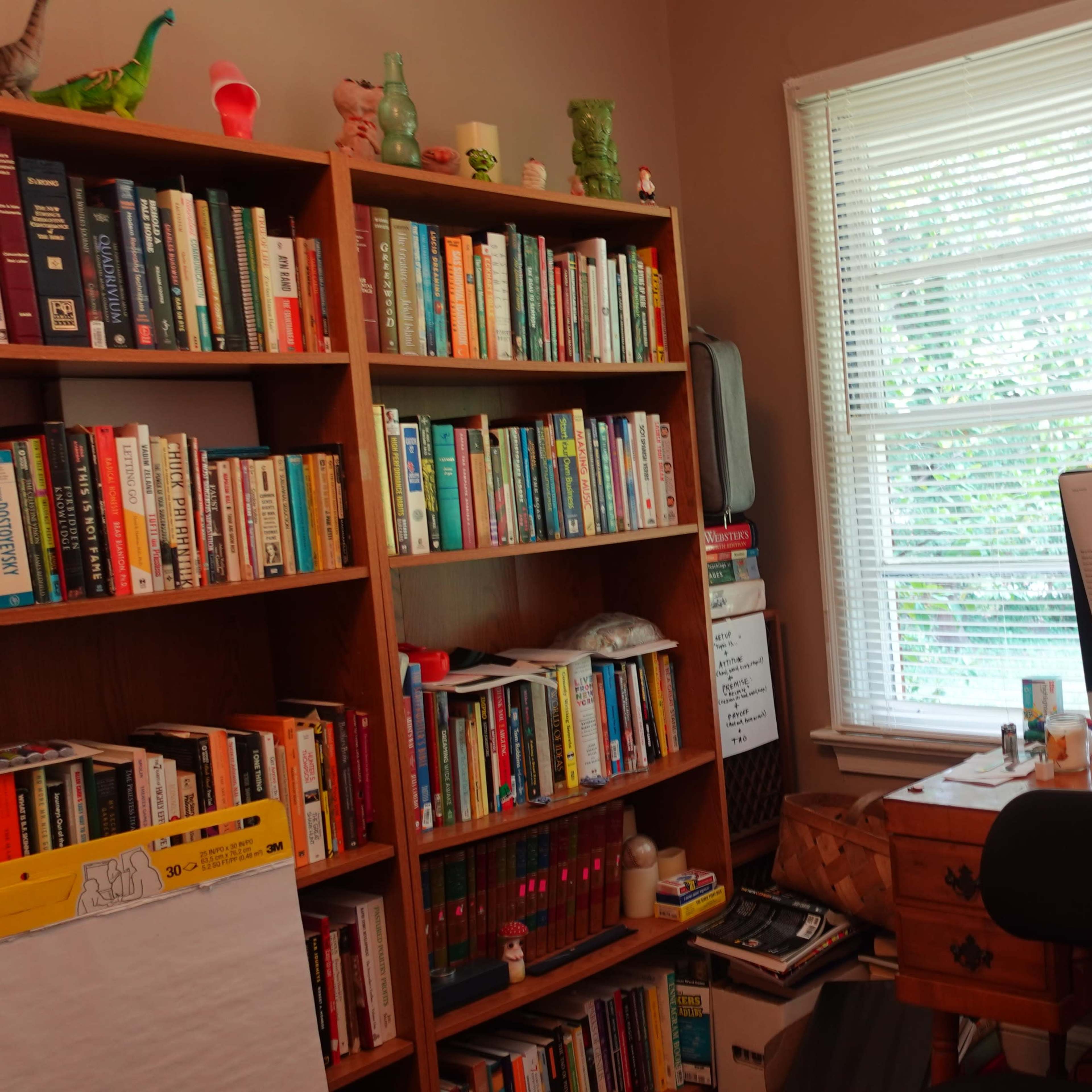 A home office features a wooden desk with a computer and stacks of papers, surrounded by bookshelves filled with books and decorative items under a window.