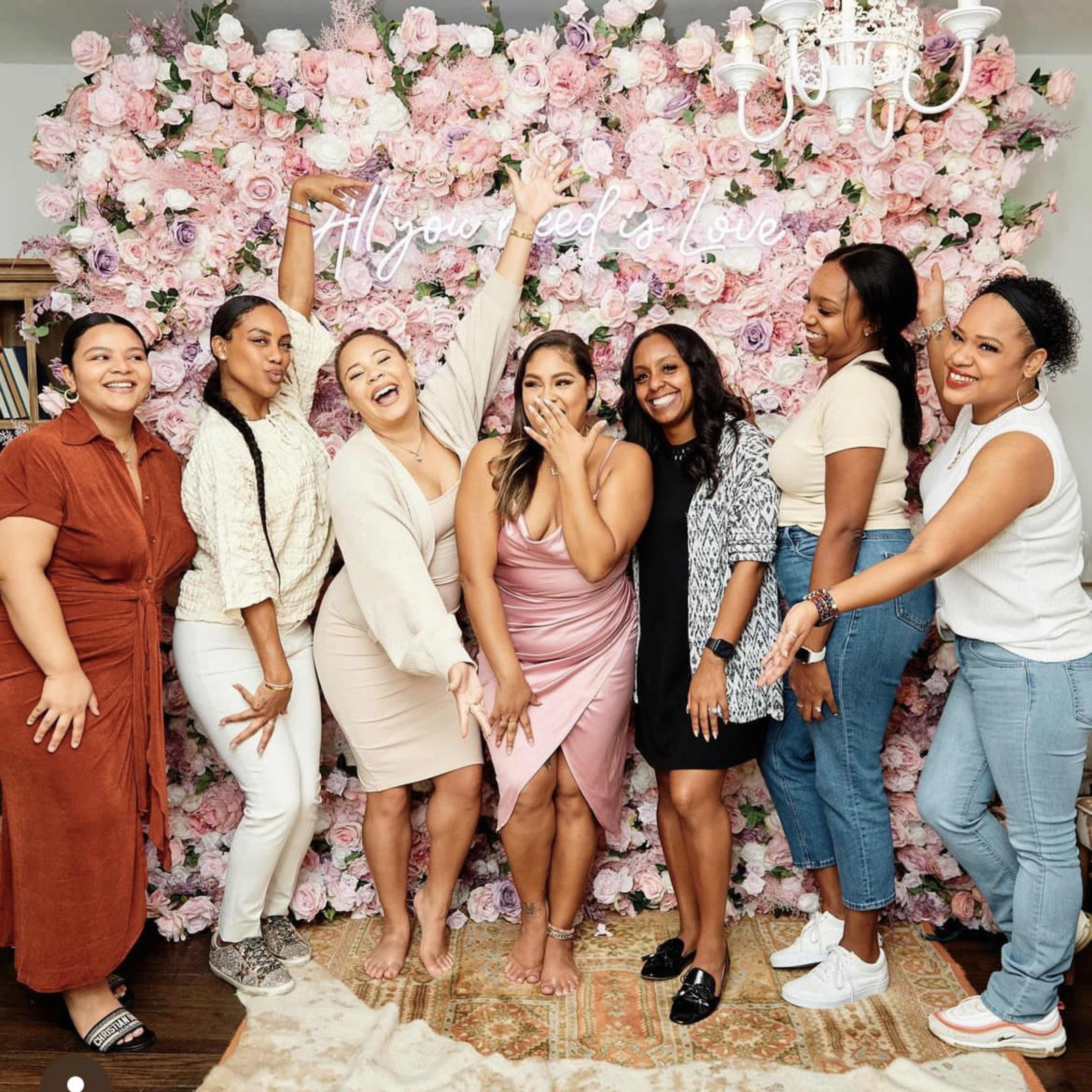 A group of women poses together in front of a wall adorned with pink flowers and a sign that reads "All you need is love."