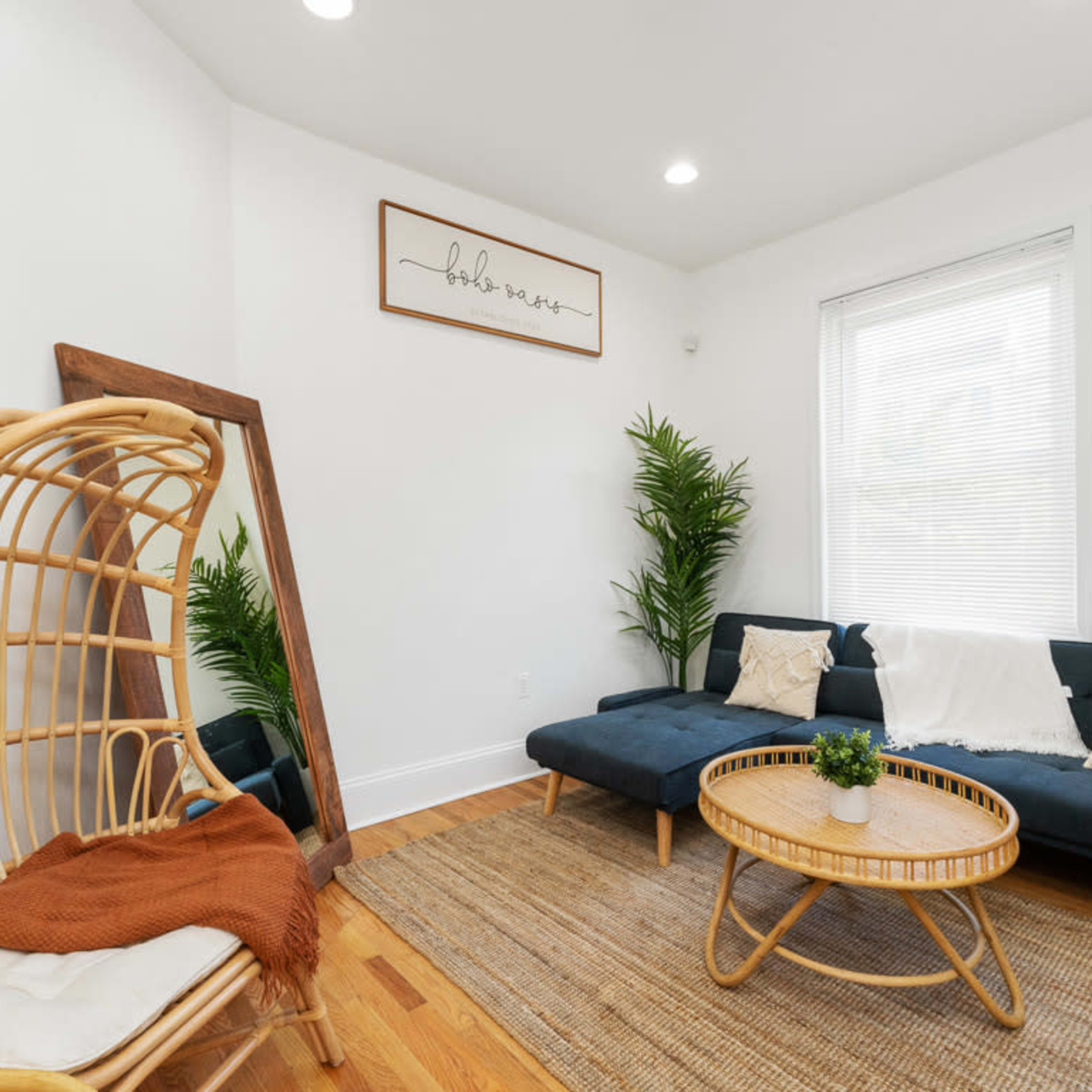 A modern living room features a dark sofa, a round coffee table, a rattan chair, and indoor plants against a light-colored wall.