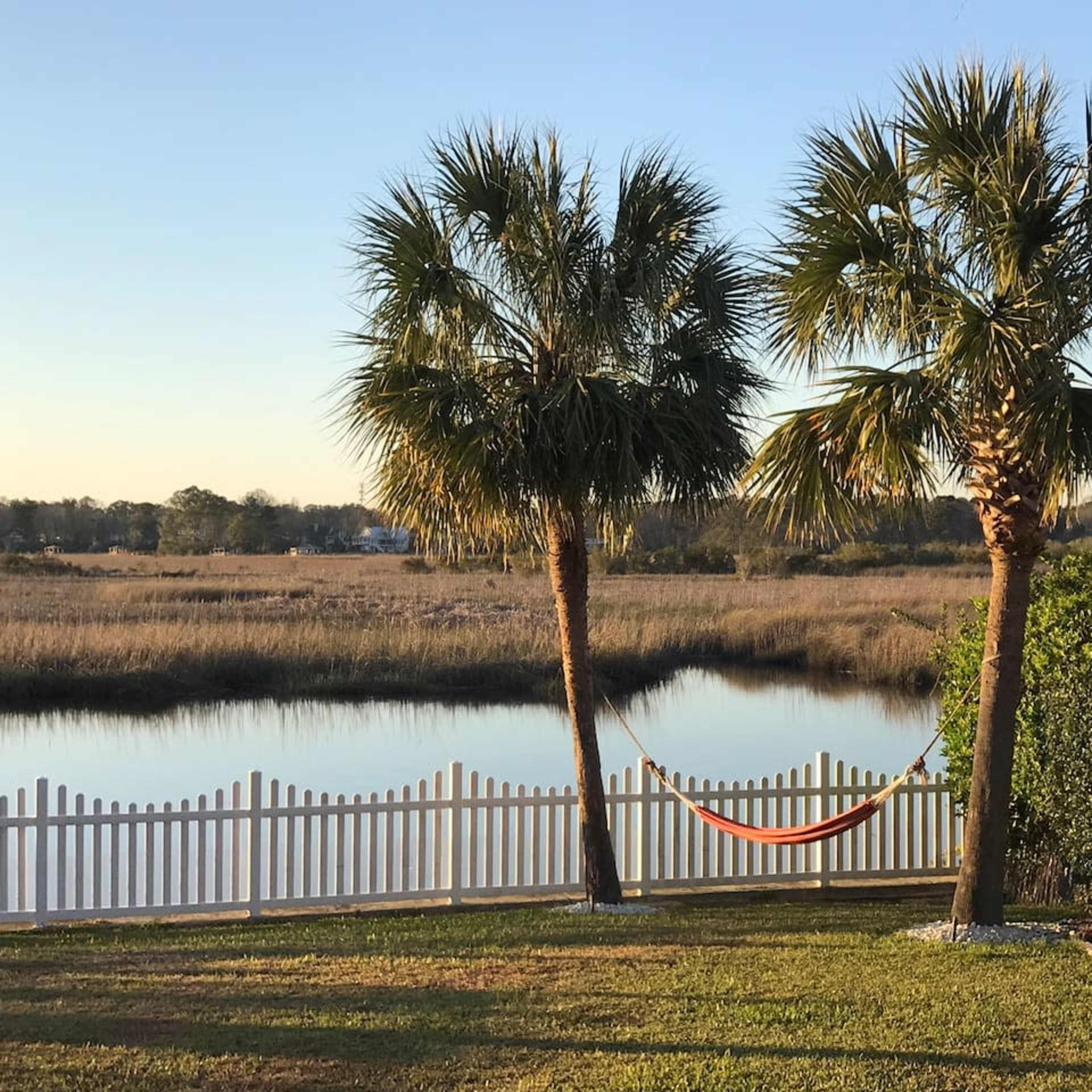 Two palm trees flank an orange hammock hanging between them, with a view of a calm body of water and a white picket fence in the background.