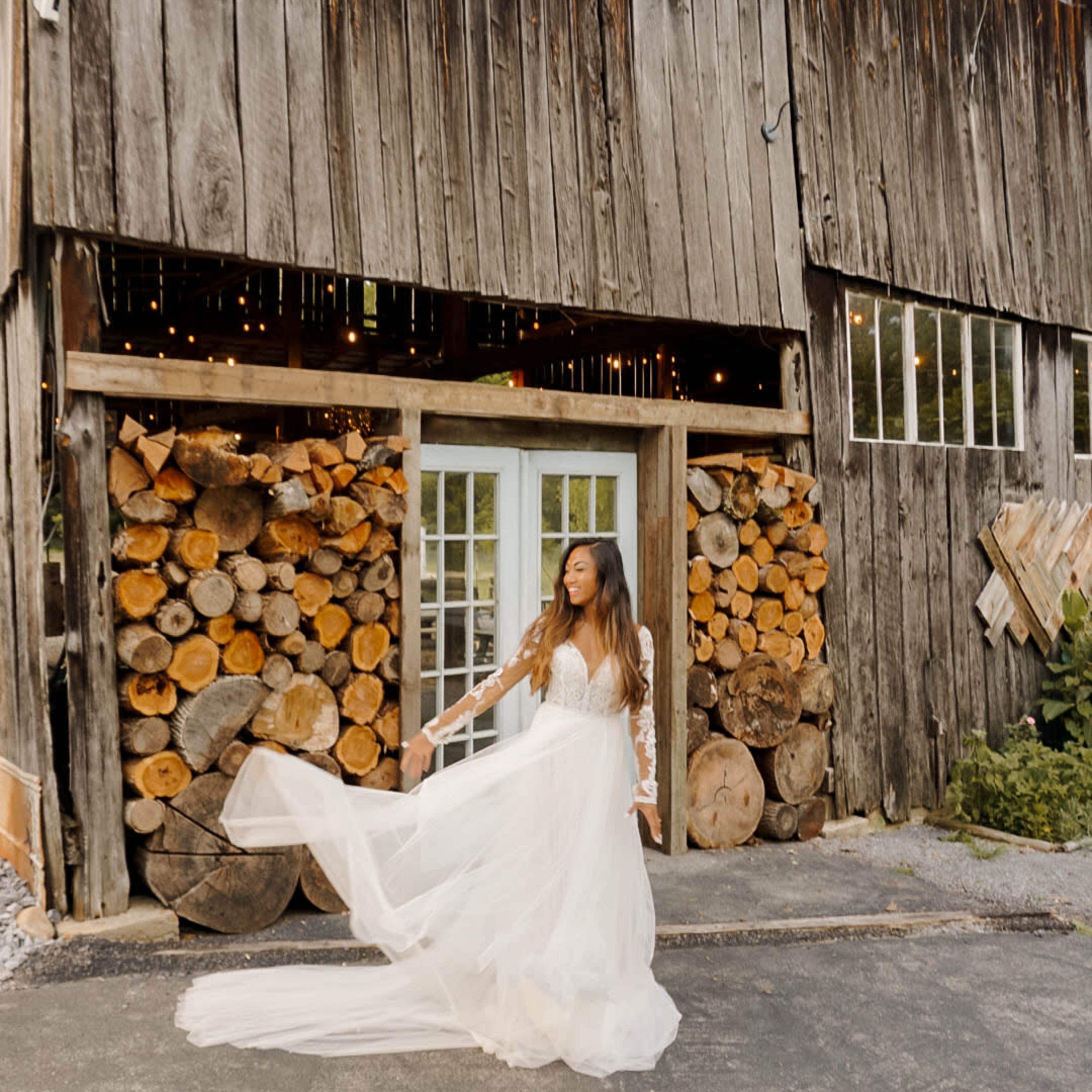 A woman in a flowing white gown poses in front of a rustic wooden barn with stacked logs.