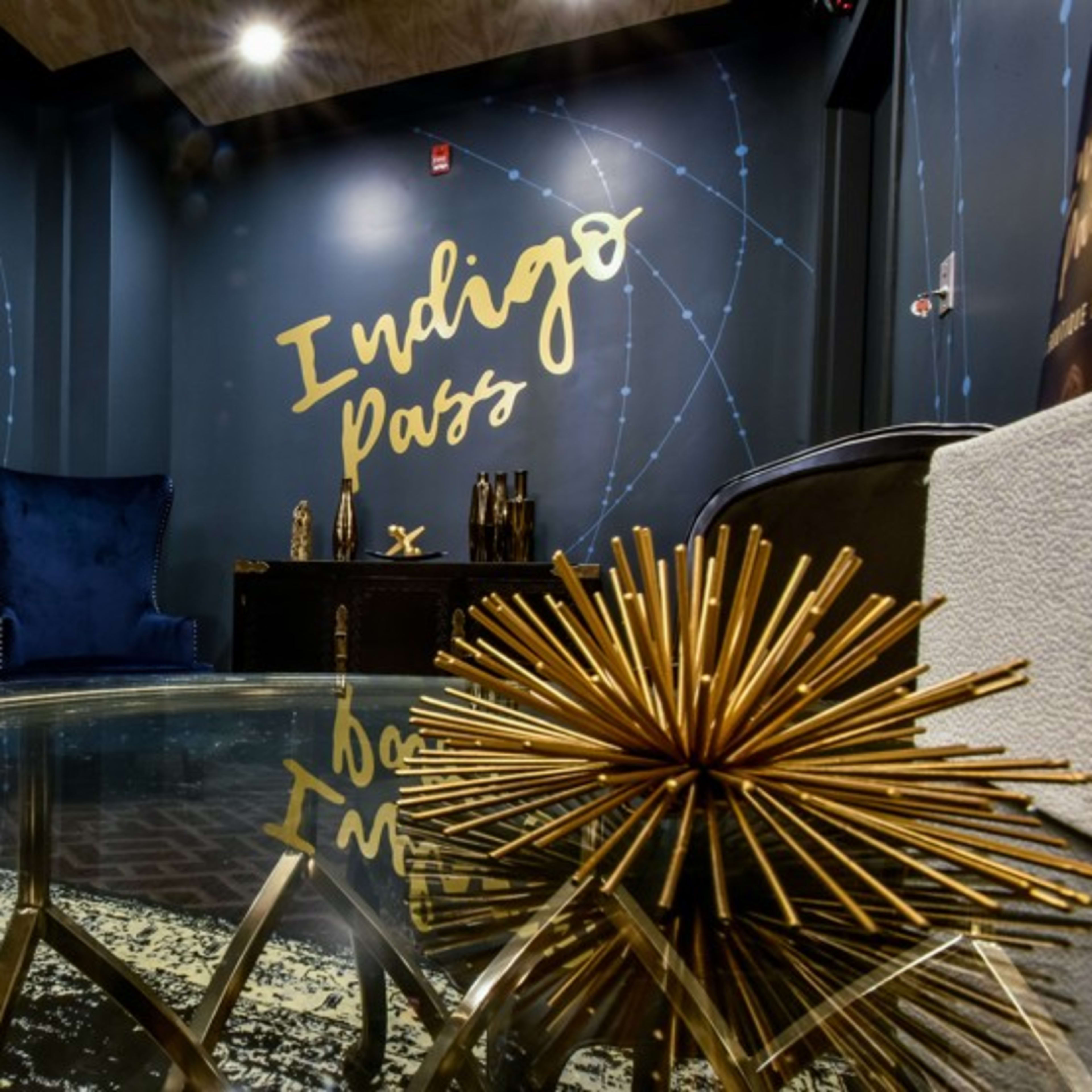 A stylish interior space with a dark blue accent wall featuring the text "Indigo Pass," a plush chair, and a decorative gold starburst object on a glass table.