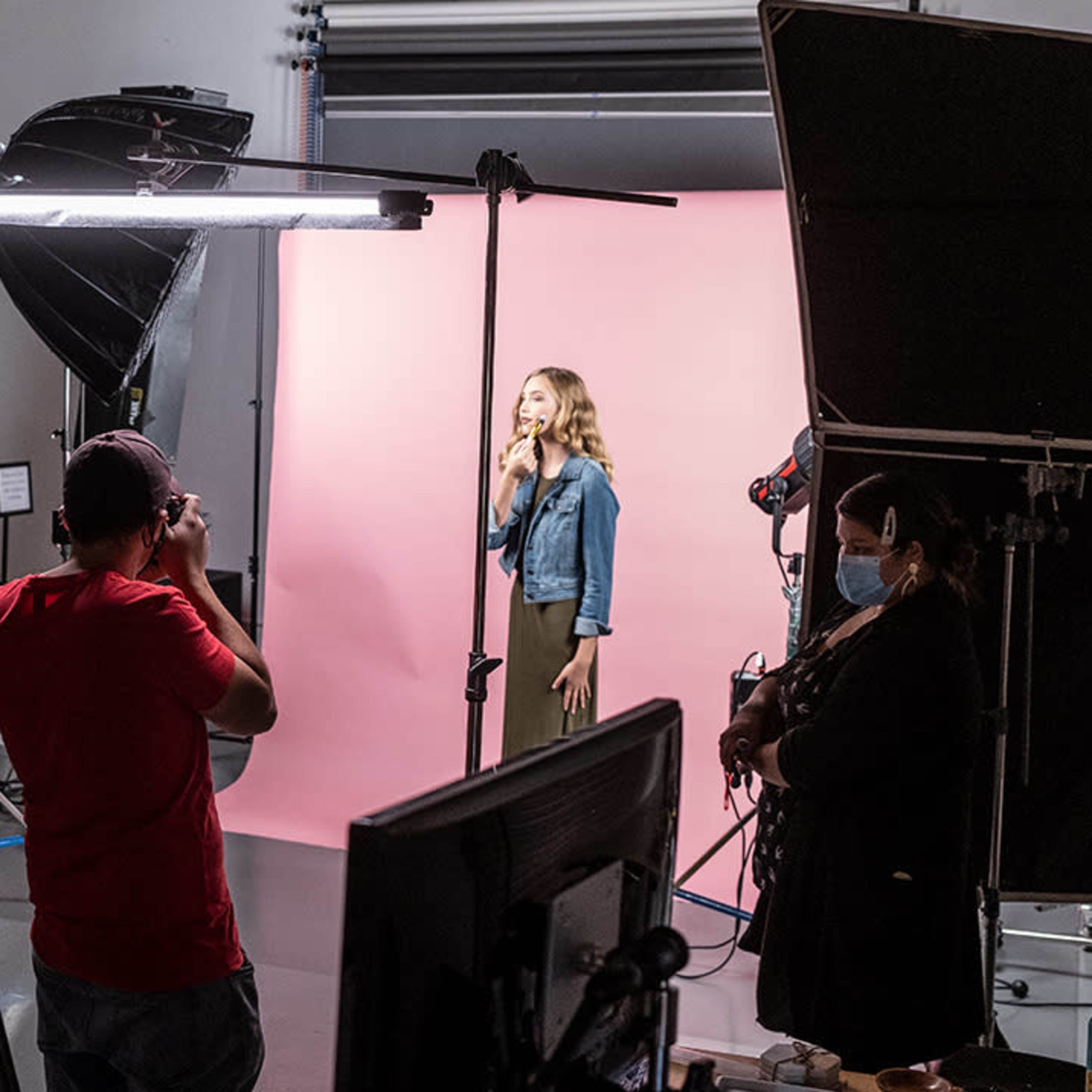 A lighting setup and filming equipment surround a model posing in front of a pink backdrop in a photography studio.