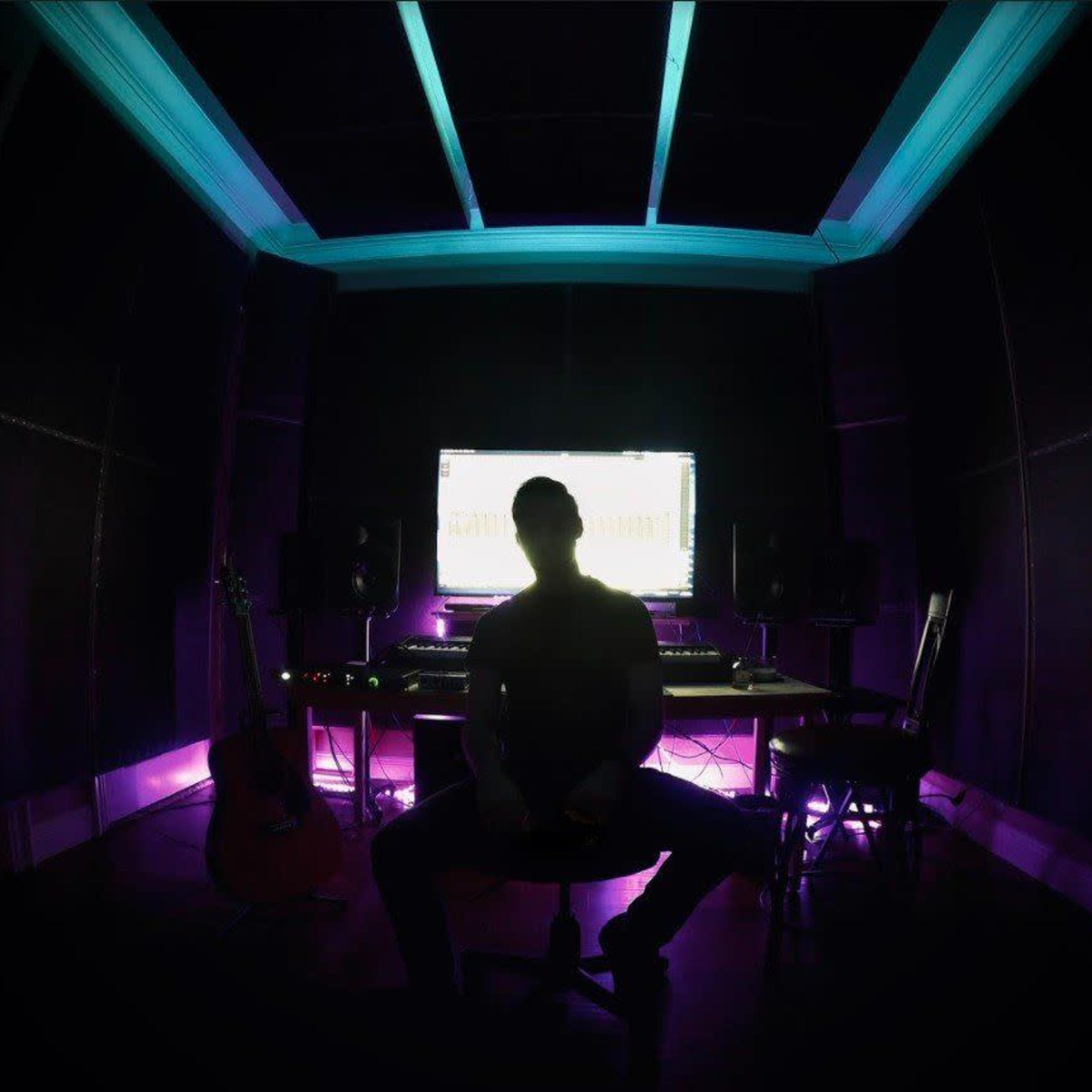 A person sits in a chair in a dimly lit music studio with a computer screen and musical instruments visible in the background.