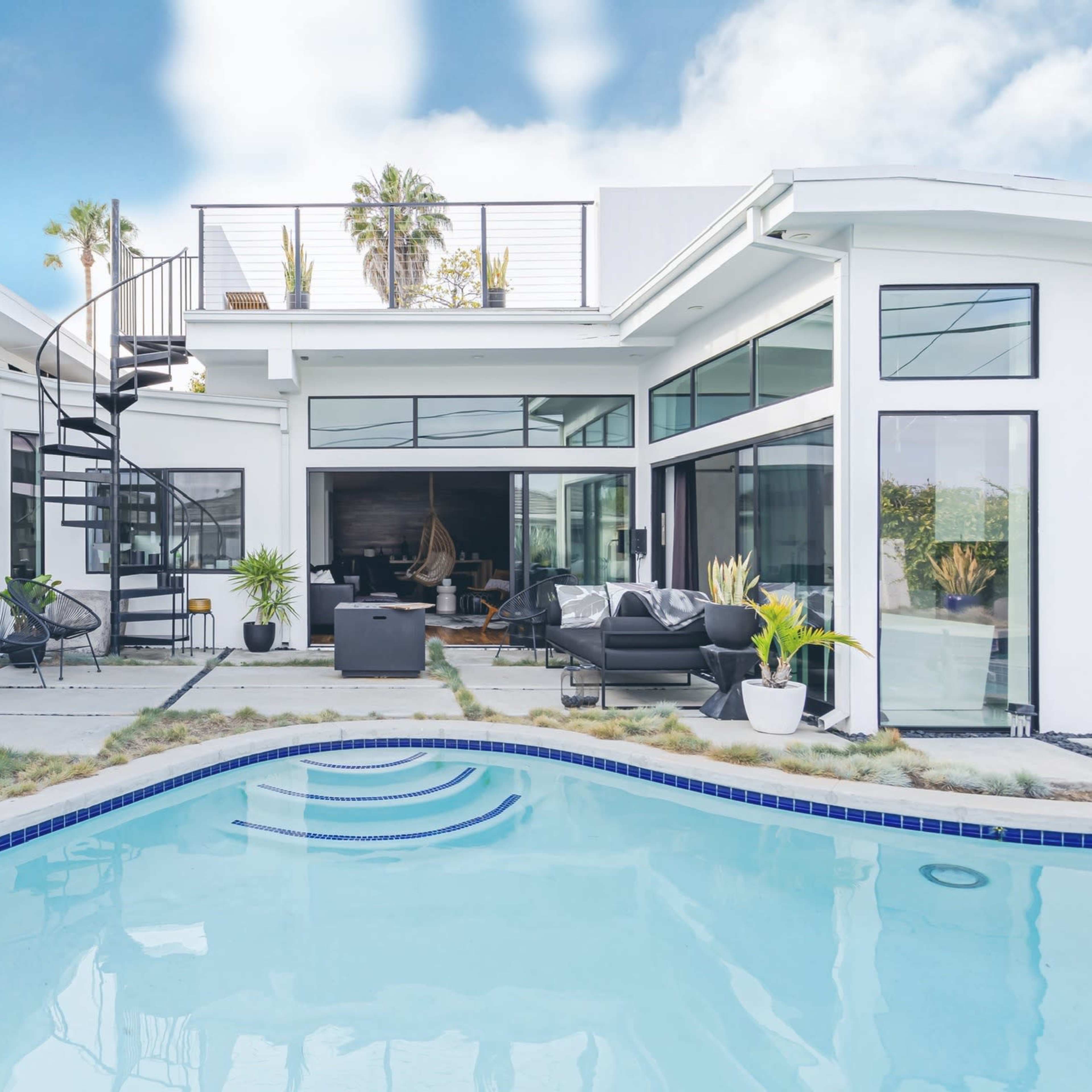 A modern house features a clean, rectangular pool surrounded by concrete patios and large windows, with a spiral staircase leading to a roof deck.