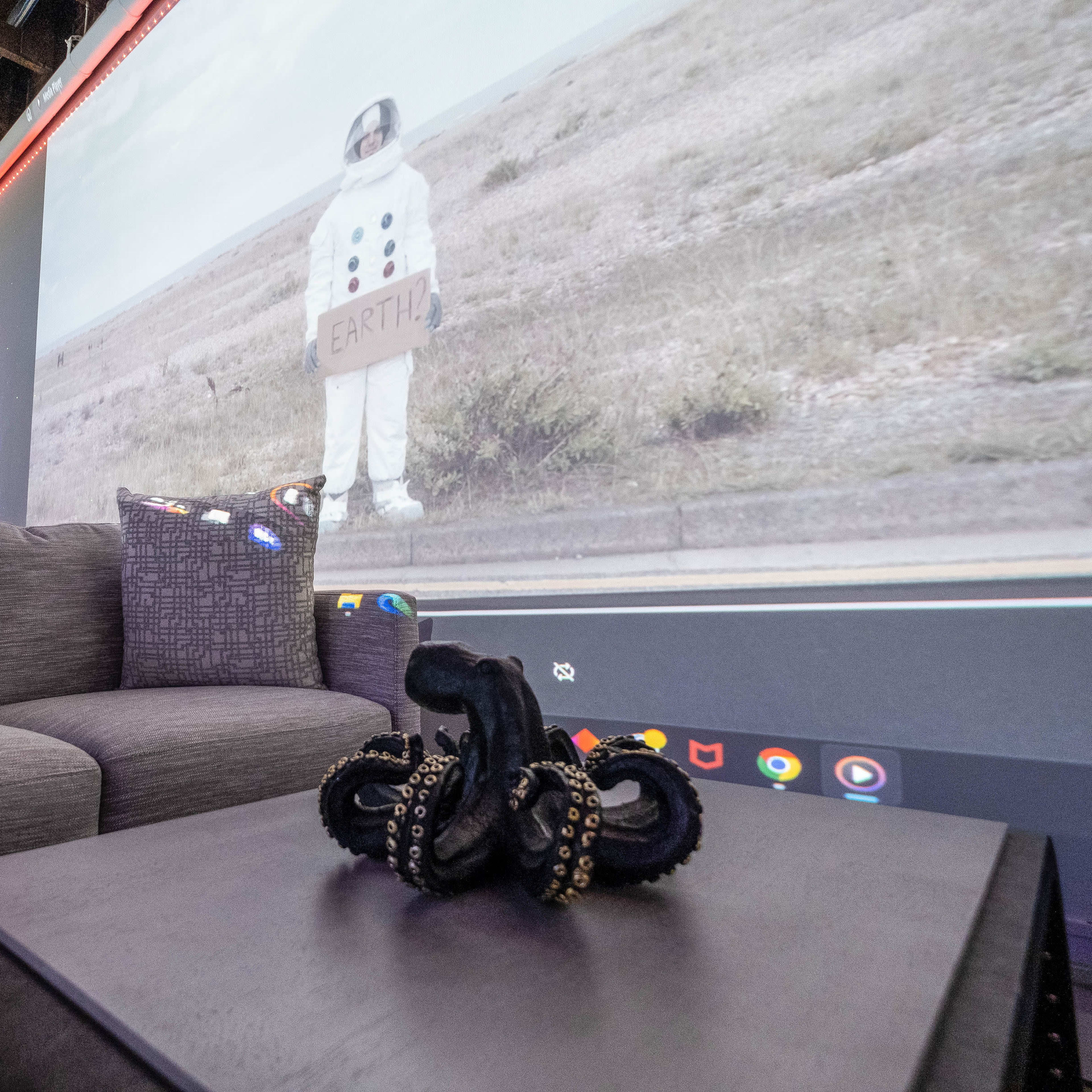 A gray couch sits in the foreground with a dark octopus sculpture on a table, while a projection of an astronaut holding a sign reading "EARTH" is visible on the wall behind.