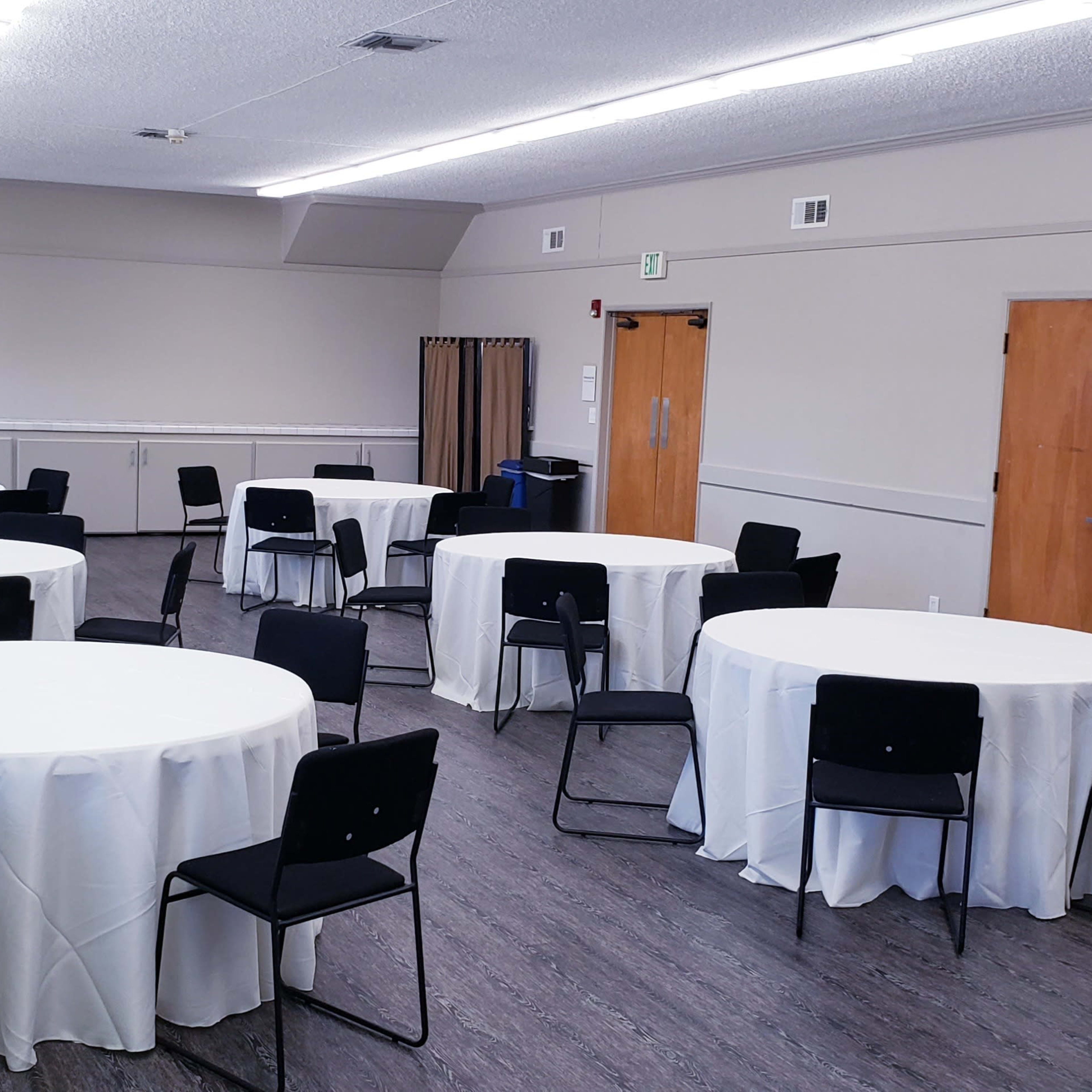 A meeting room with eight round tables covered in white tablecloths, each surrounded by black chairs.