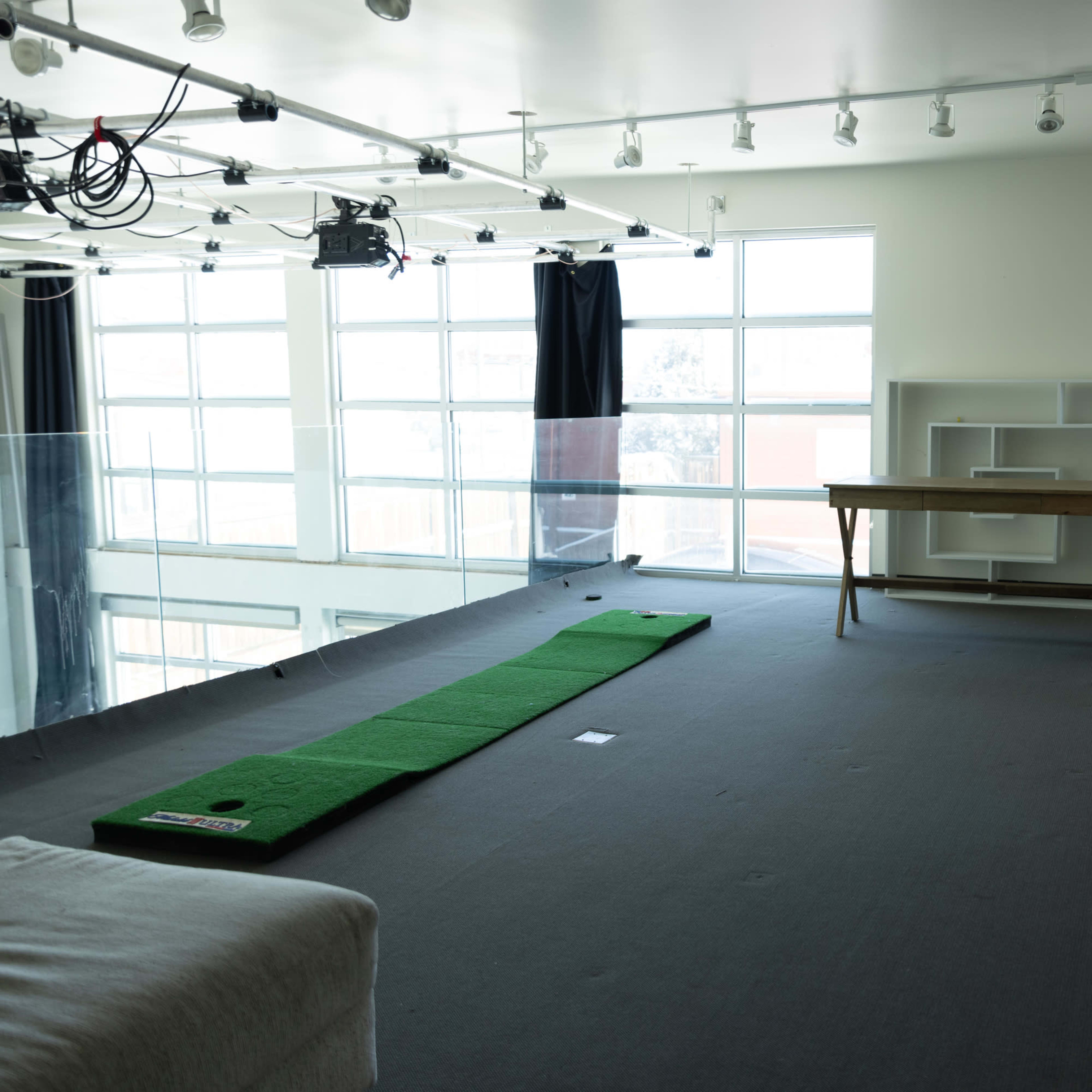 A spacious, brightly lit room with large windows, featuring a putting green on the floor, a wooden table, and a sideboard.