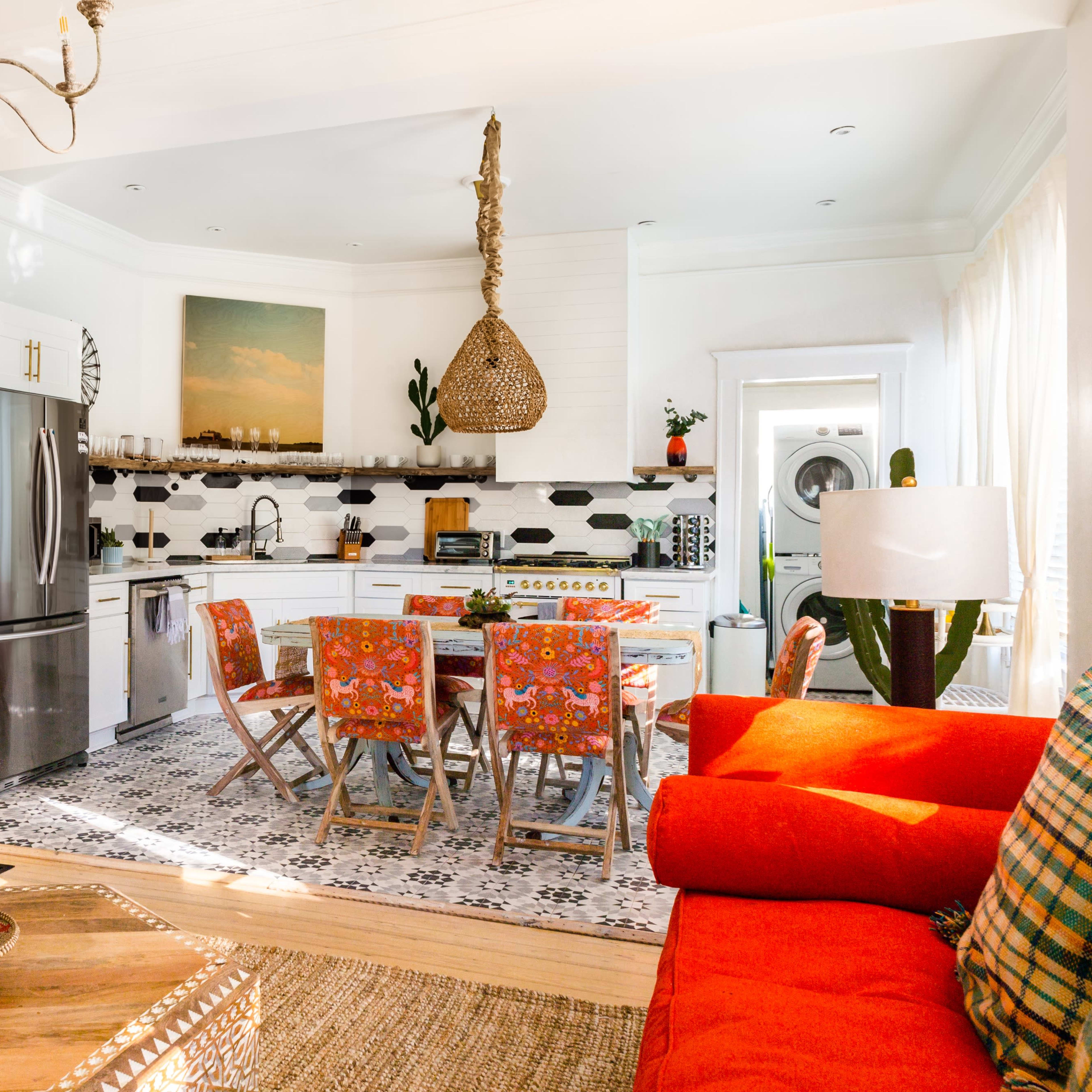 A bright, colorful kitchen and living area feature a dining table surrounded by patterned chairs, with a modern refrigerator and decorative elements like plants and rugs.