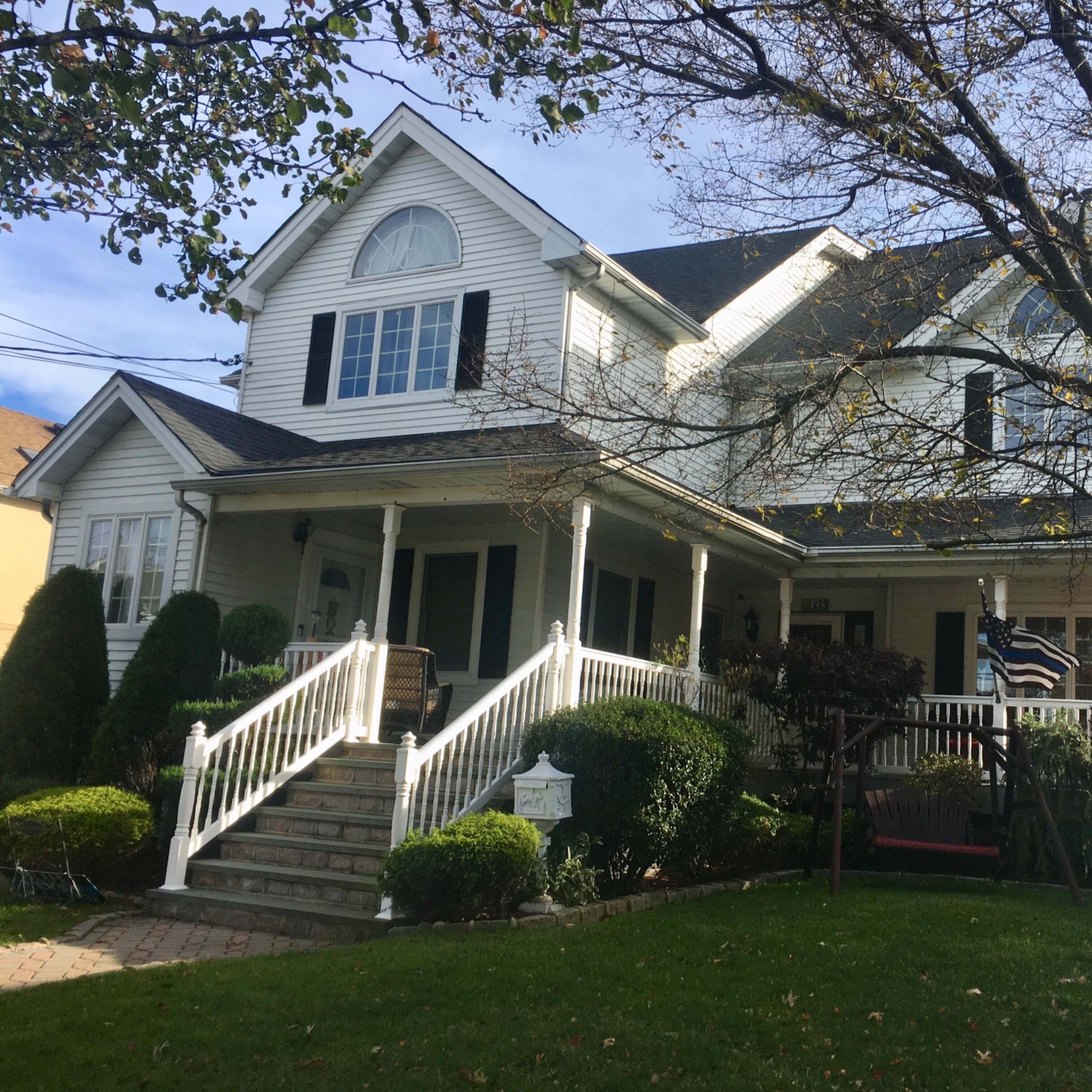 A large two-story house with a porch, surrounded by neatly trimmed shrubs and trees, features white siding and multiple gabled roofs.