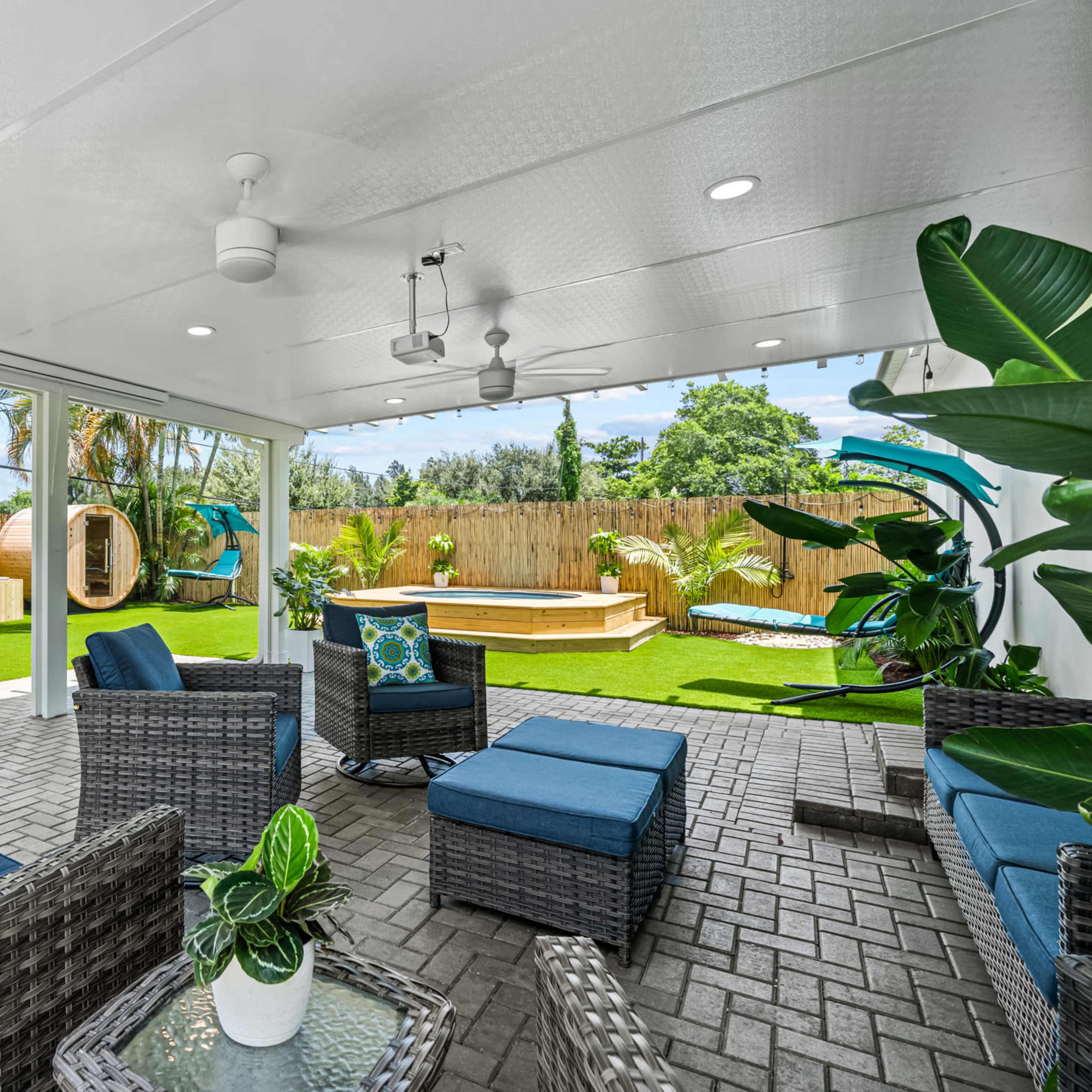 A patio area with wicker furniture, a small round hot tub, and a green yard surrounded by palm trees and a wooden fence.