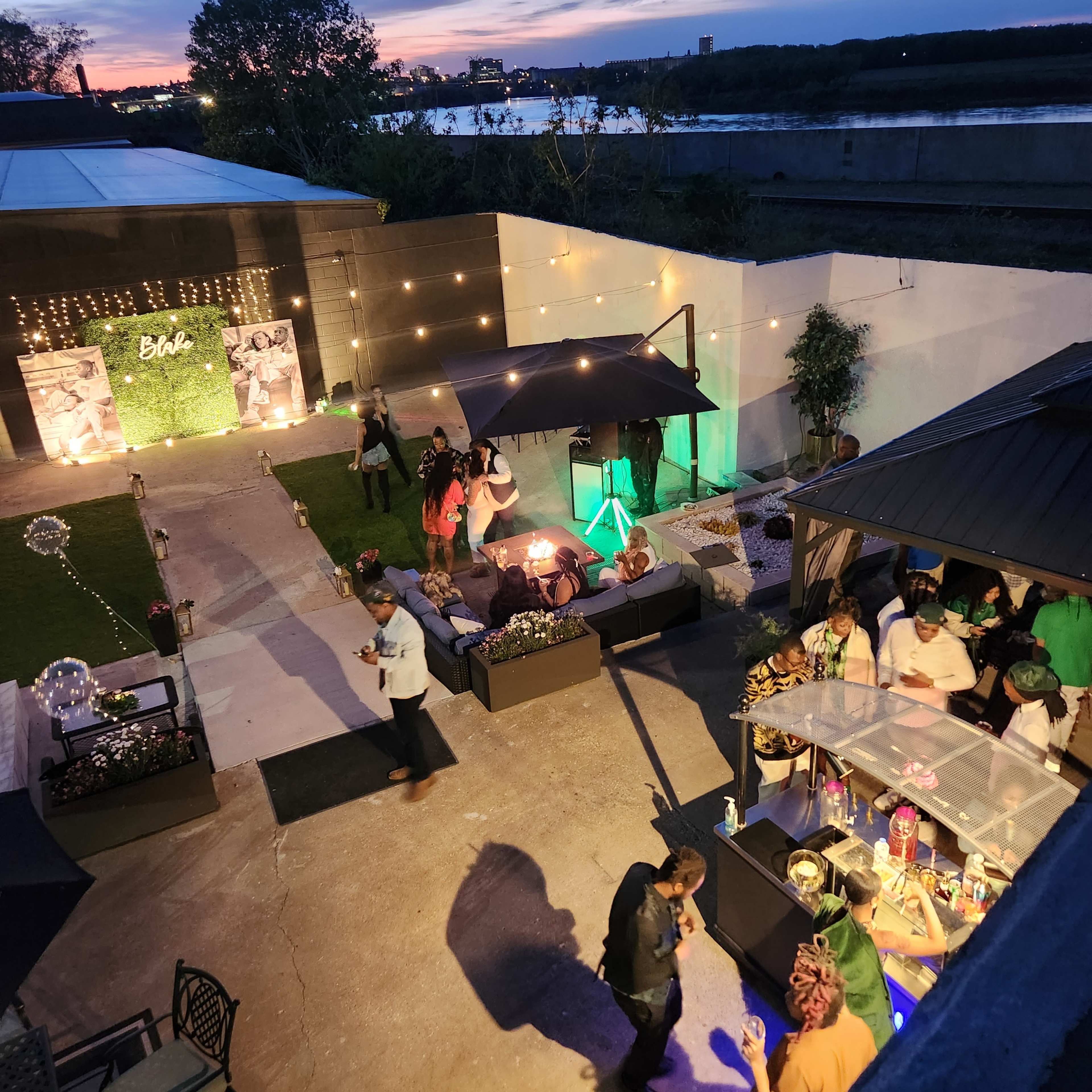 The scene features an outdoor event space with guests mingling under string lights, surrounded by greenery, and a bar area in one corner.