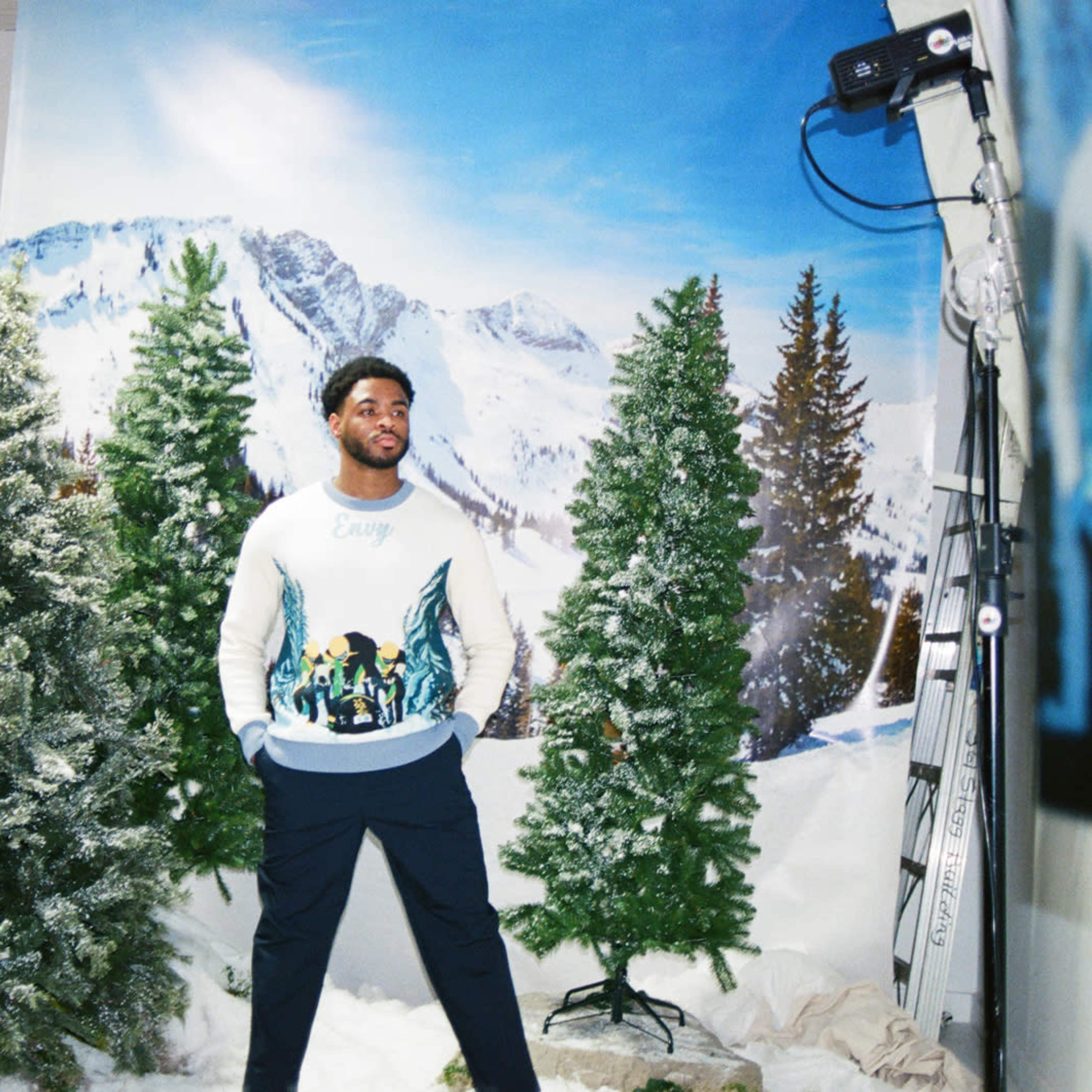 A person stands in front of a painted winter landscape backdrop surrounded by artificial trees.
