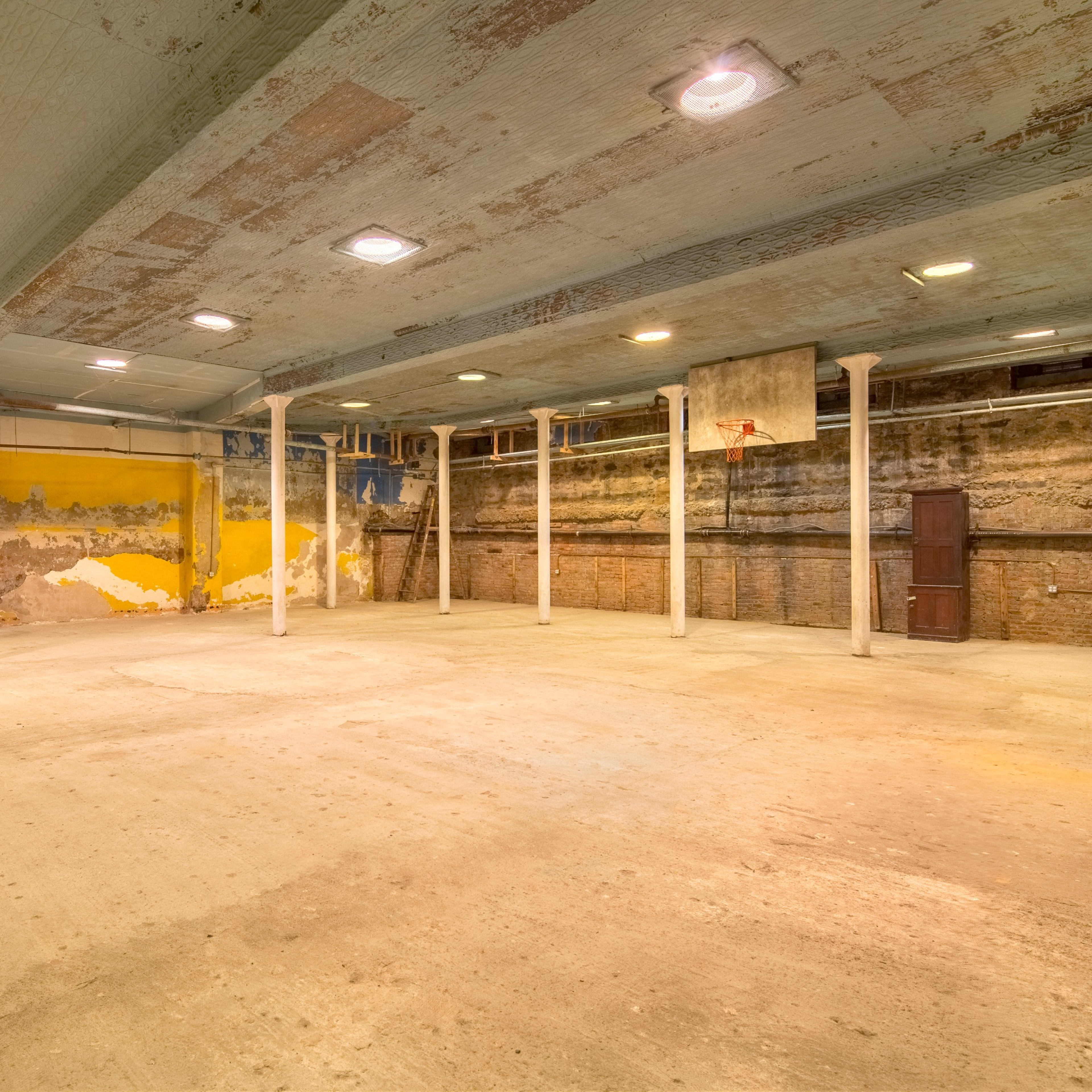 An empty indoor basketball court with worn walls, a basketball hoop mounted on a backboard, and exposed concrete flooring.