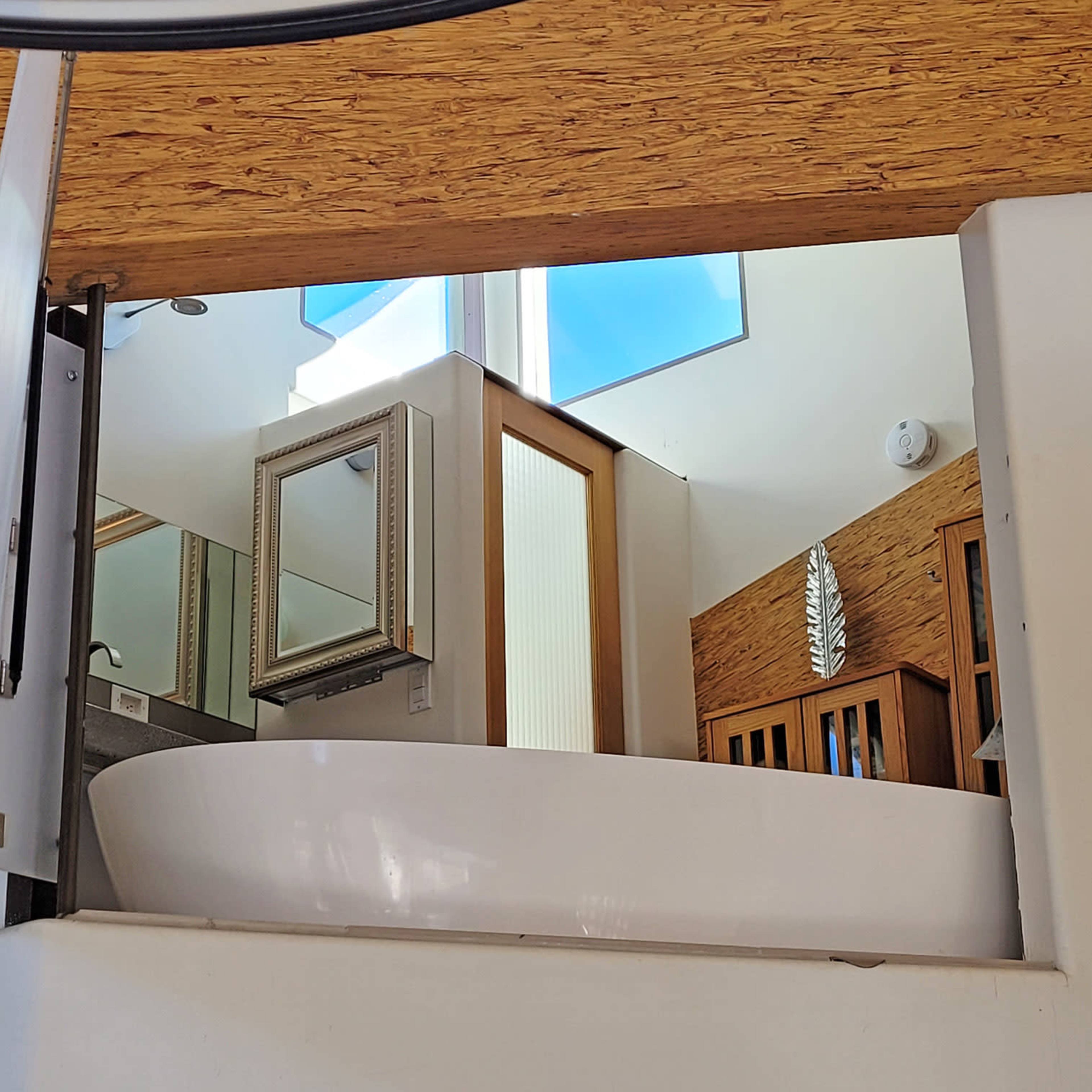 A view of a modern bathroom with a freestanding bathtub, a mirror, and wooden cabinetry, framed by a sloped ceiling and large windows.