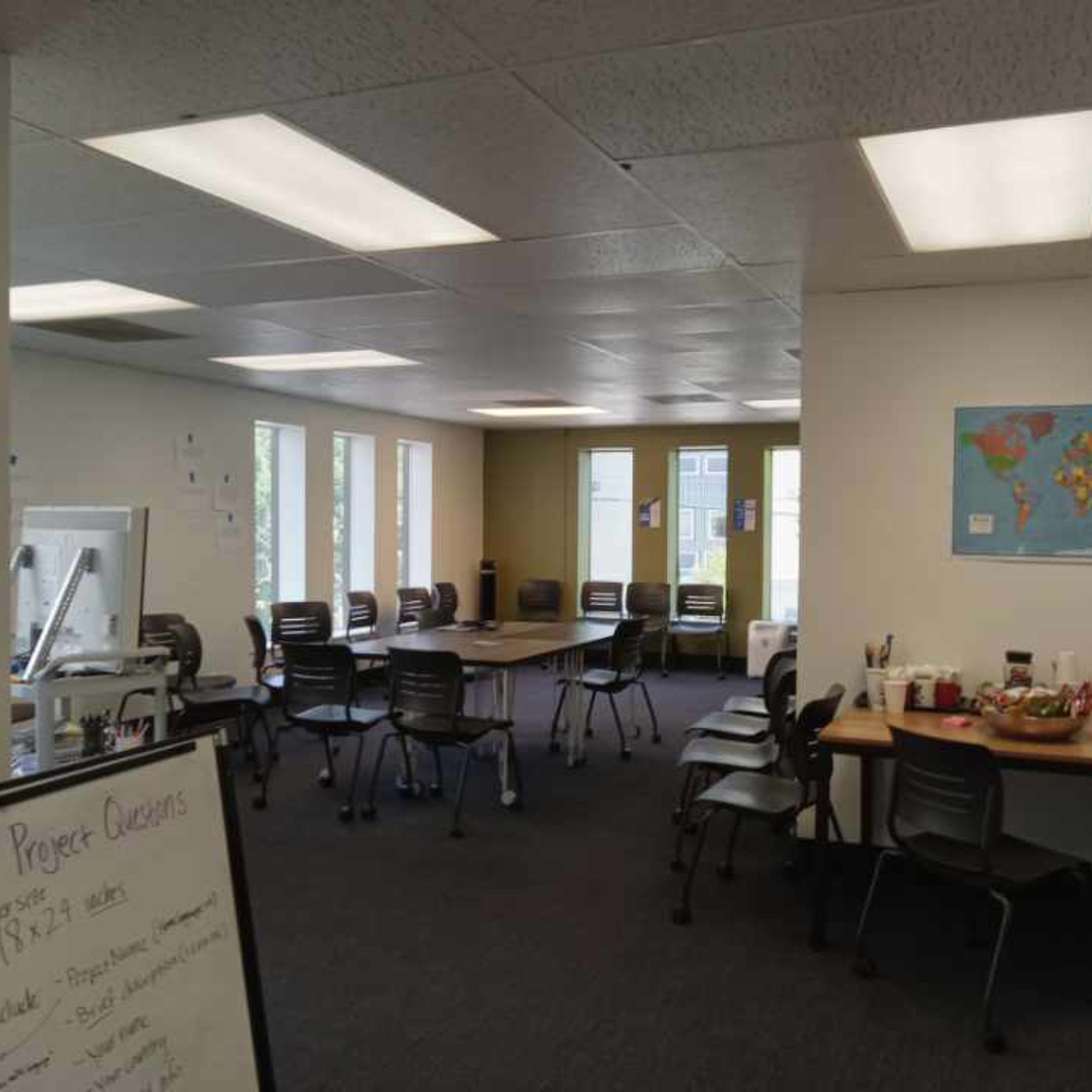 A spacious office with several tables and chairs arranged for a meeting, along with a world map on the wall and a whiteboard displaying project questions.