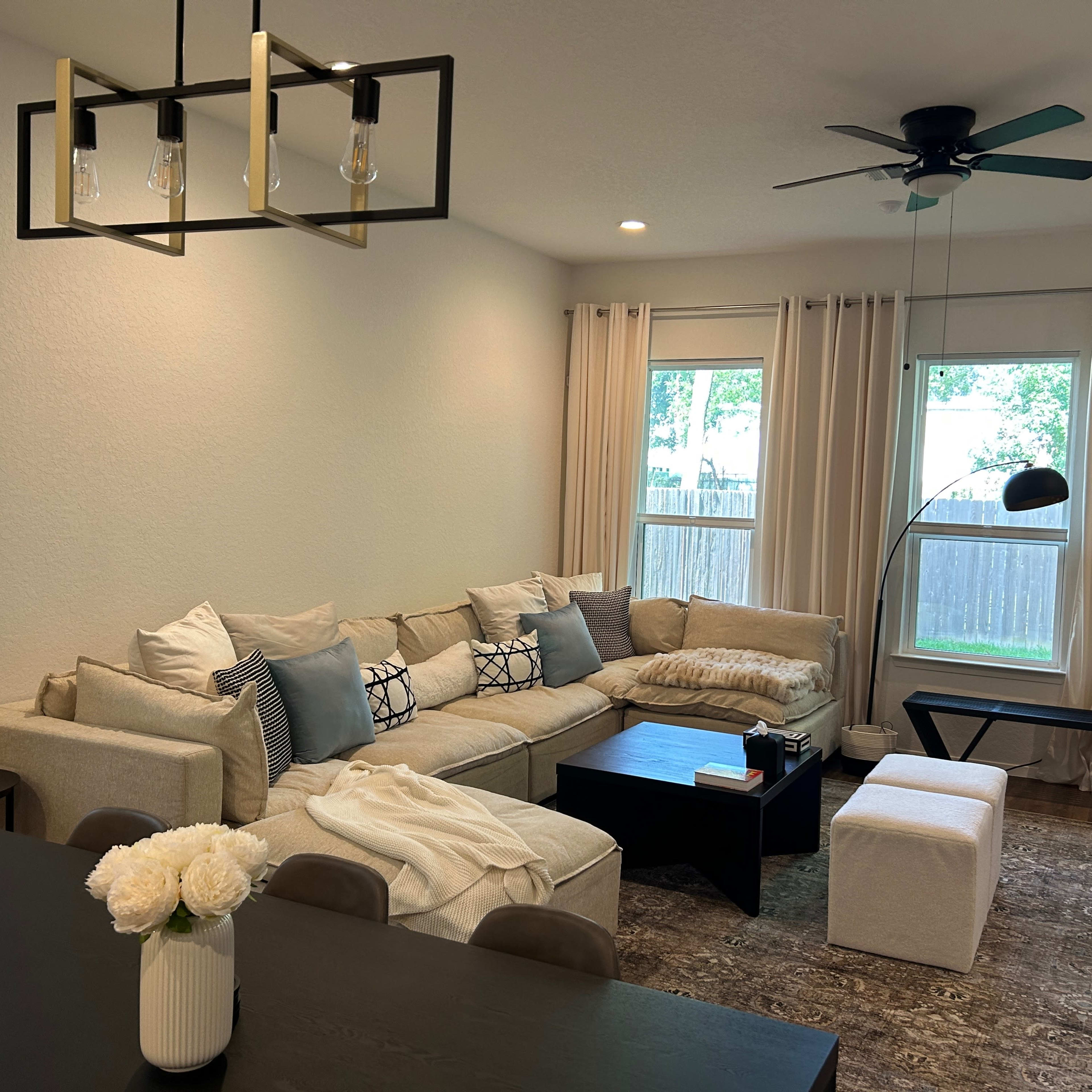 A modern living room with a large sectional sofa, a coffee table in front, and a dining area with a table and upholstered stools, all set against a backdrop of large windows.