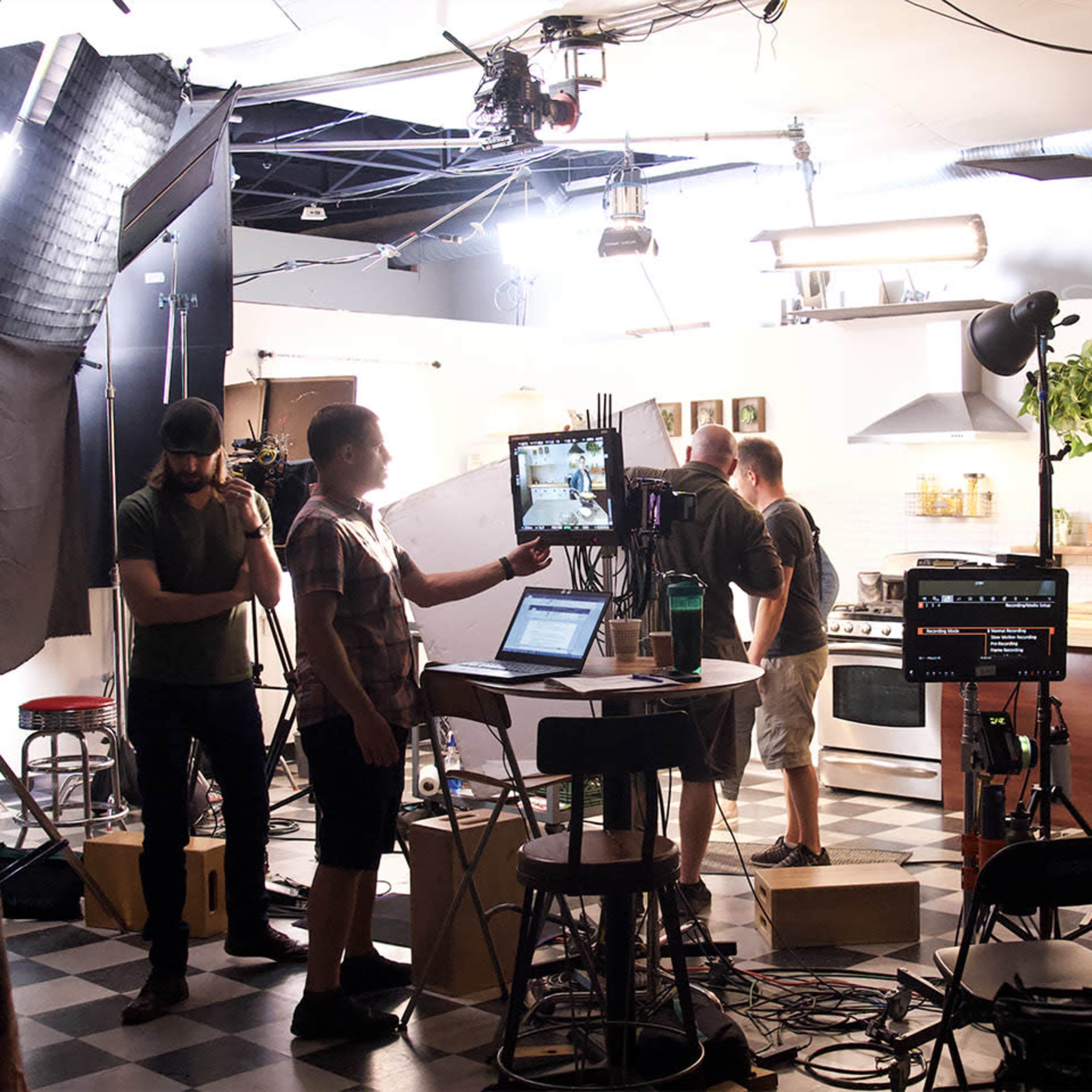 A film crew is working in a kitchen set, with various cameras and lighting equipment arranged for a shoot.