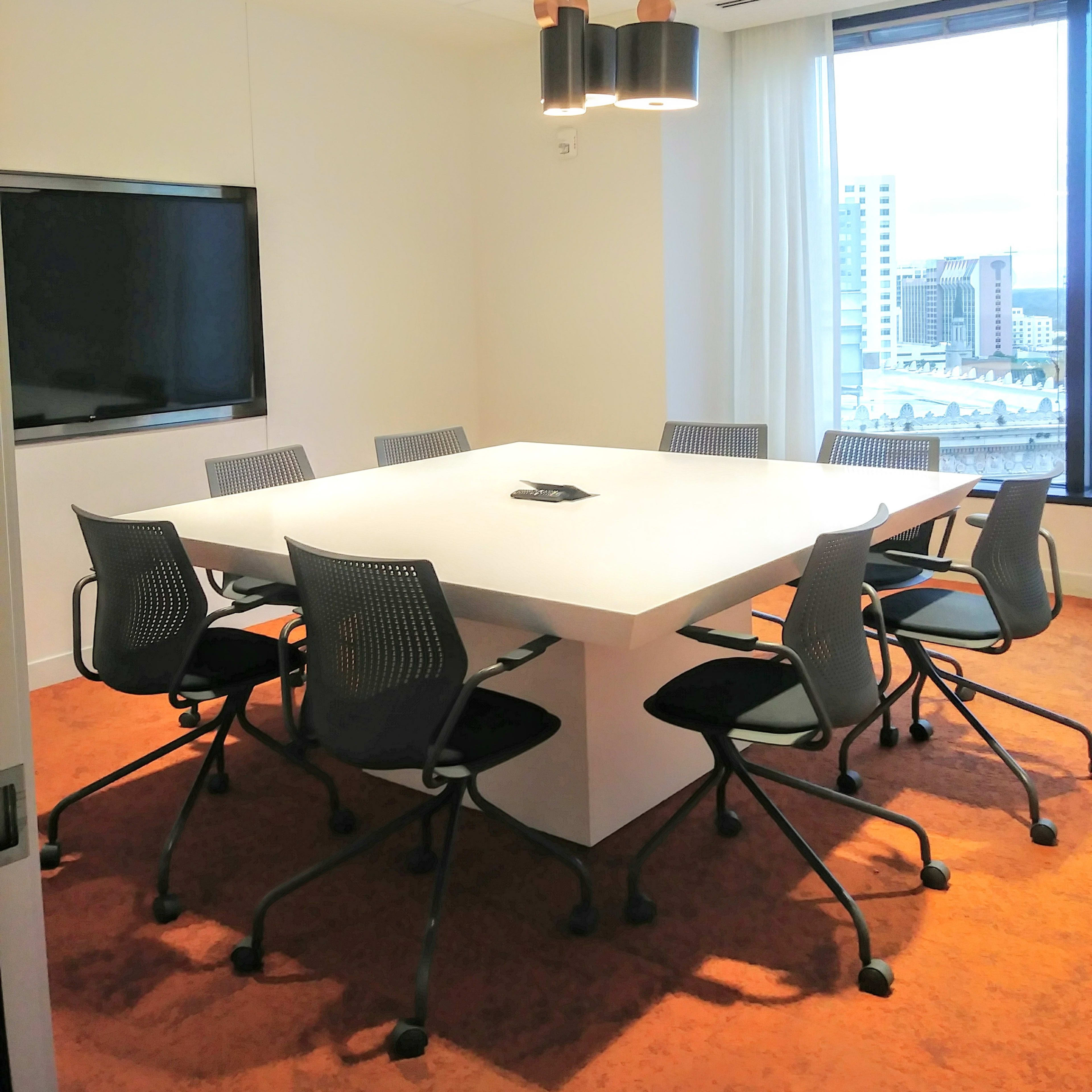 A modern conference room features a large white table surrounded by eight ergonomic chairs, with a television mounted on the wall and large windows providing a view of the city.