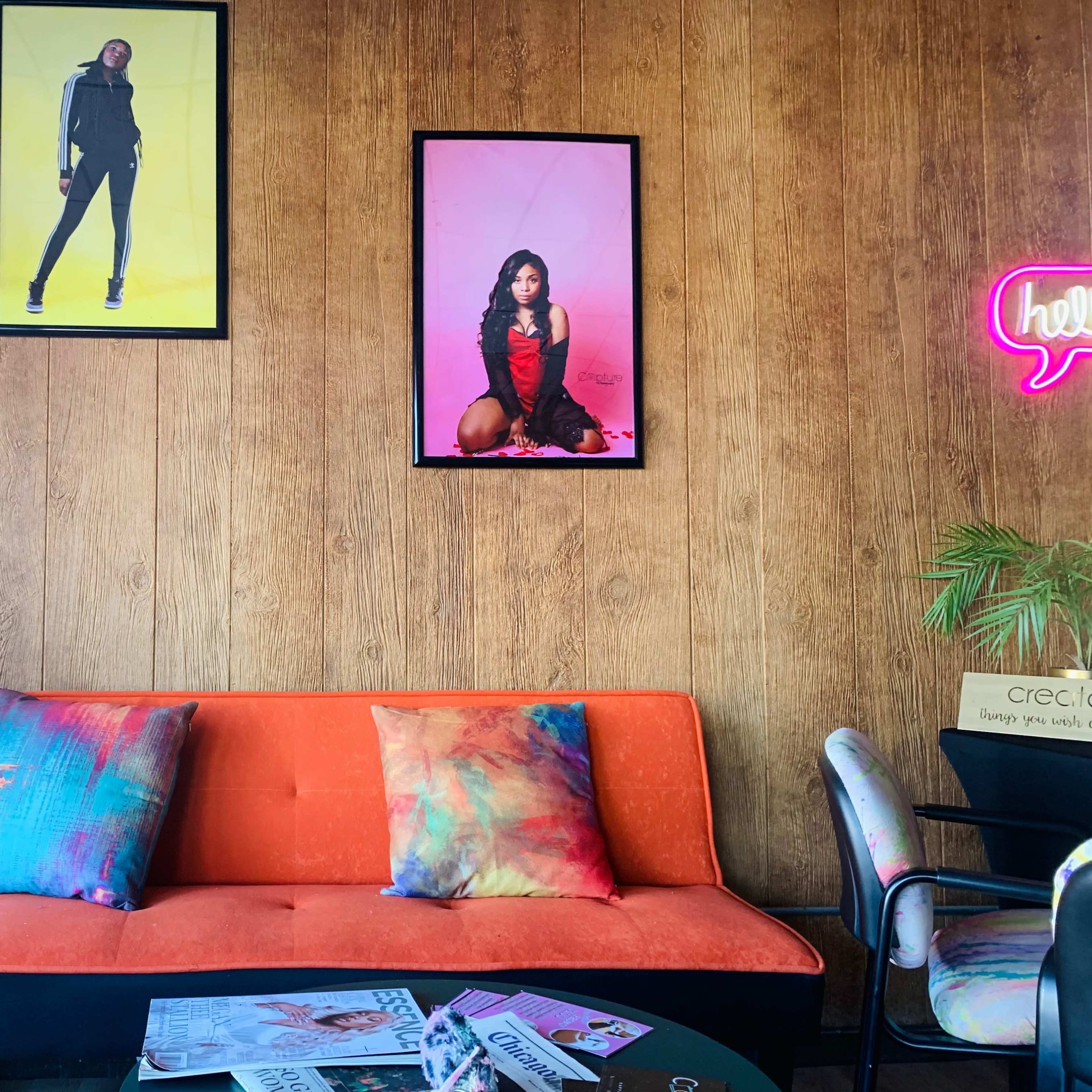 The scene features an orange sofa with colorful pillows, flanked by two framed fashion photographs on a wooden wall, alongside plants and a neon sign.