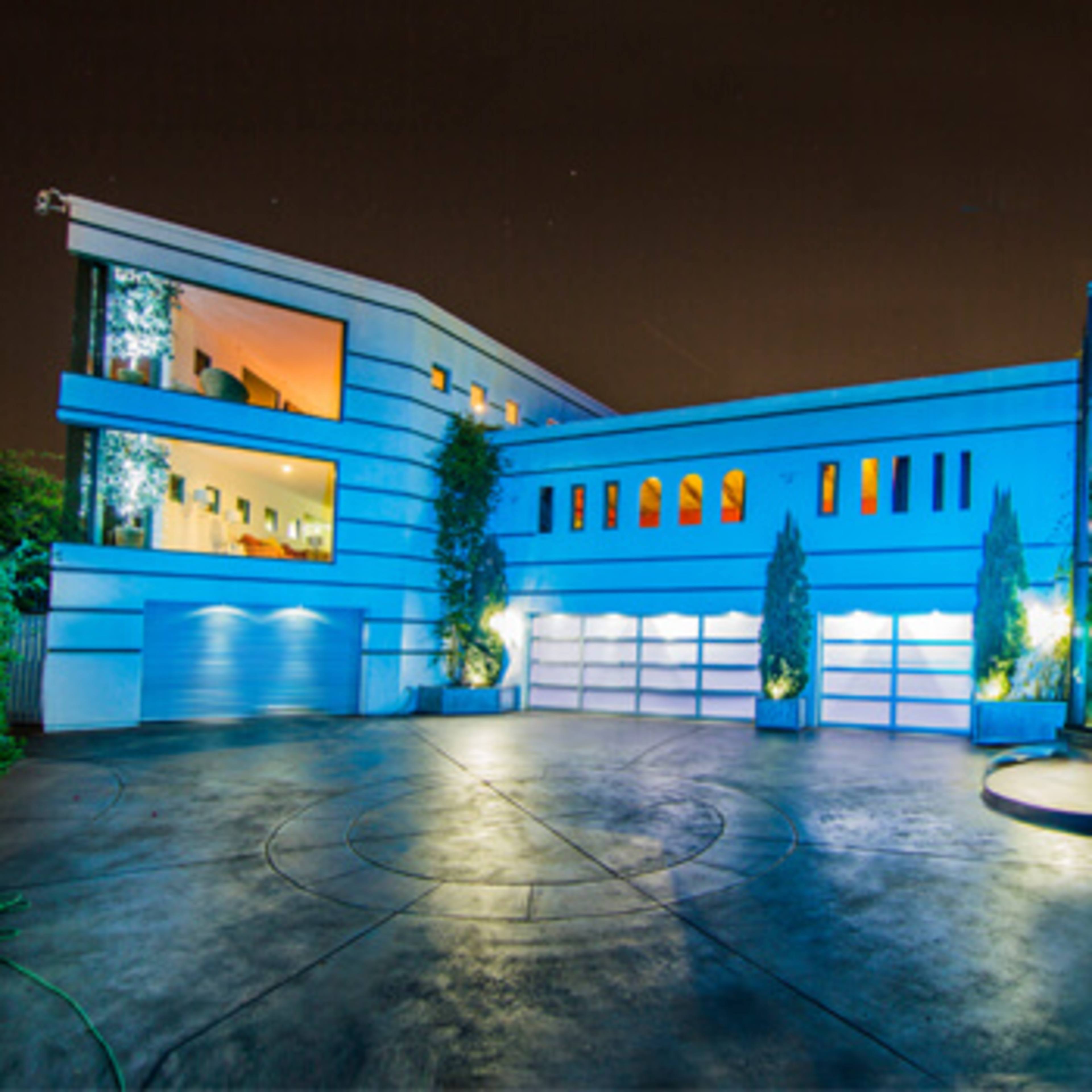A modern, two-story house features a glass-fronted entryway and is illuminated in blue at night, surrounded by landscaped greenery and a curved driveway.