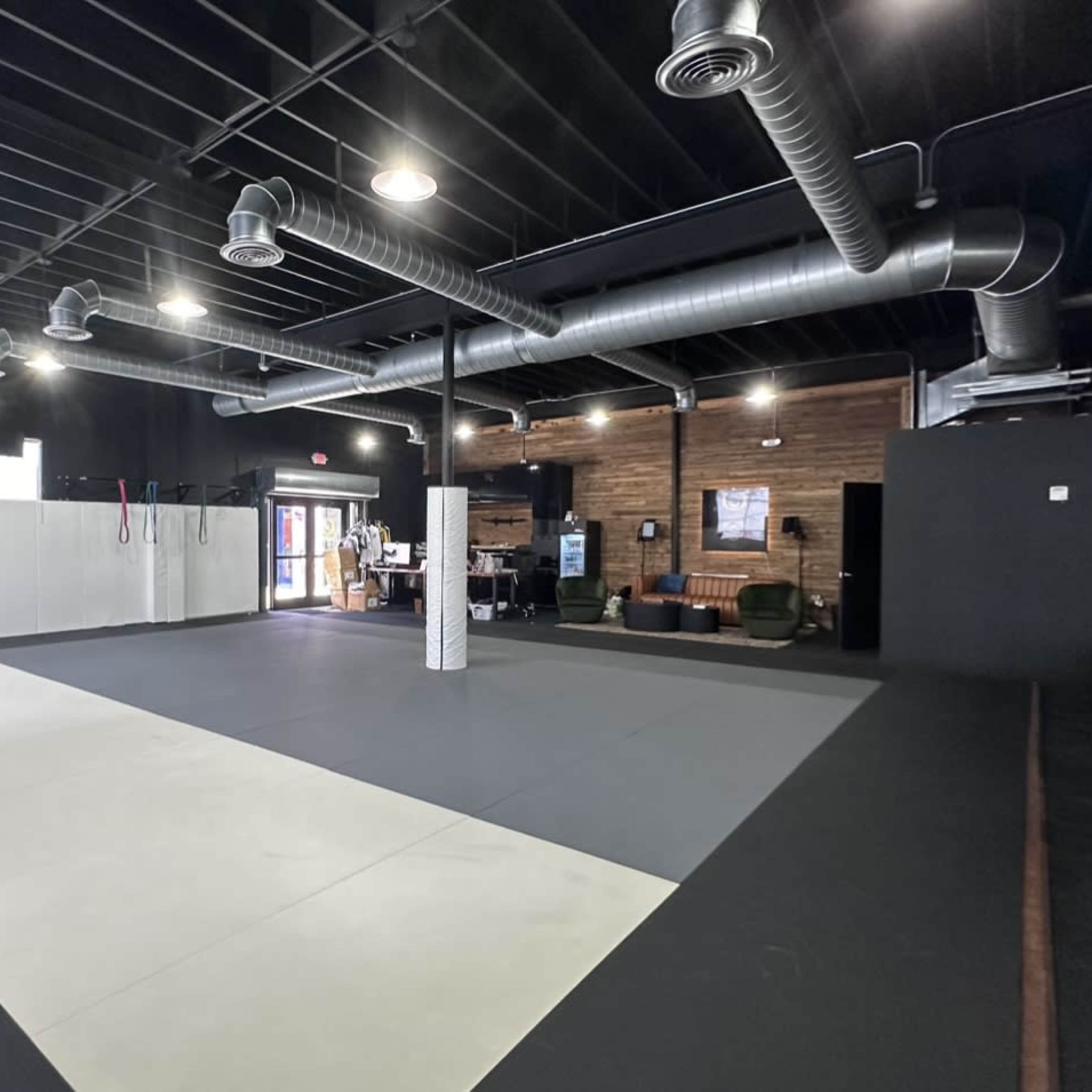 An empty training facility with a large mat area, exposed beams, and industrial-style ventilation ducts.