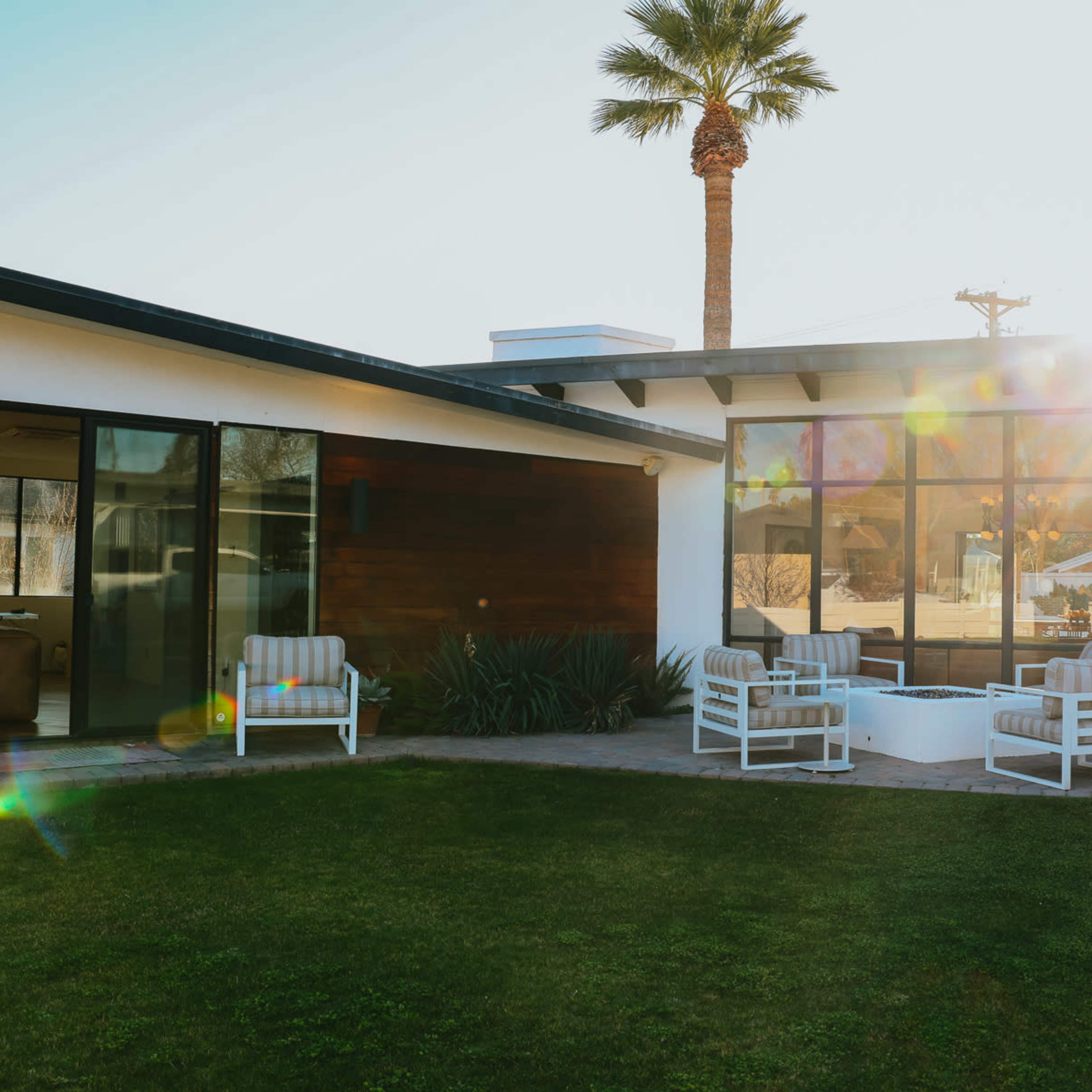 A modern, single-story house with large glass windows and a private outdoor seating area surrounded by a well-maintained lawn.
