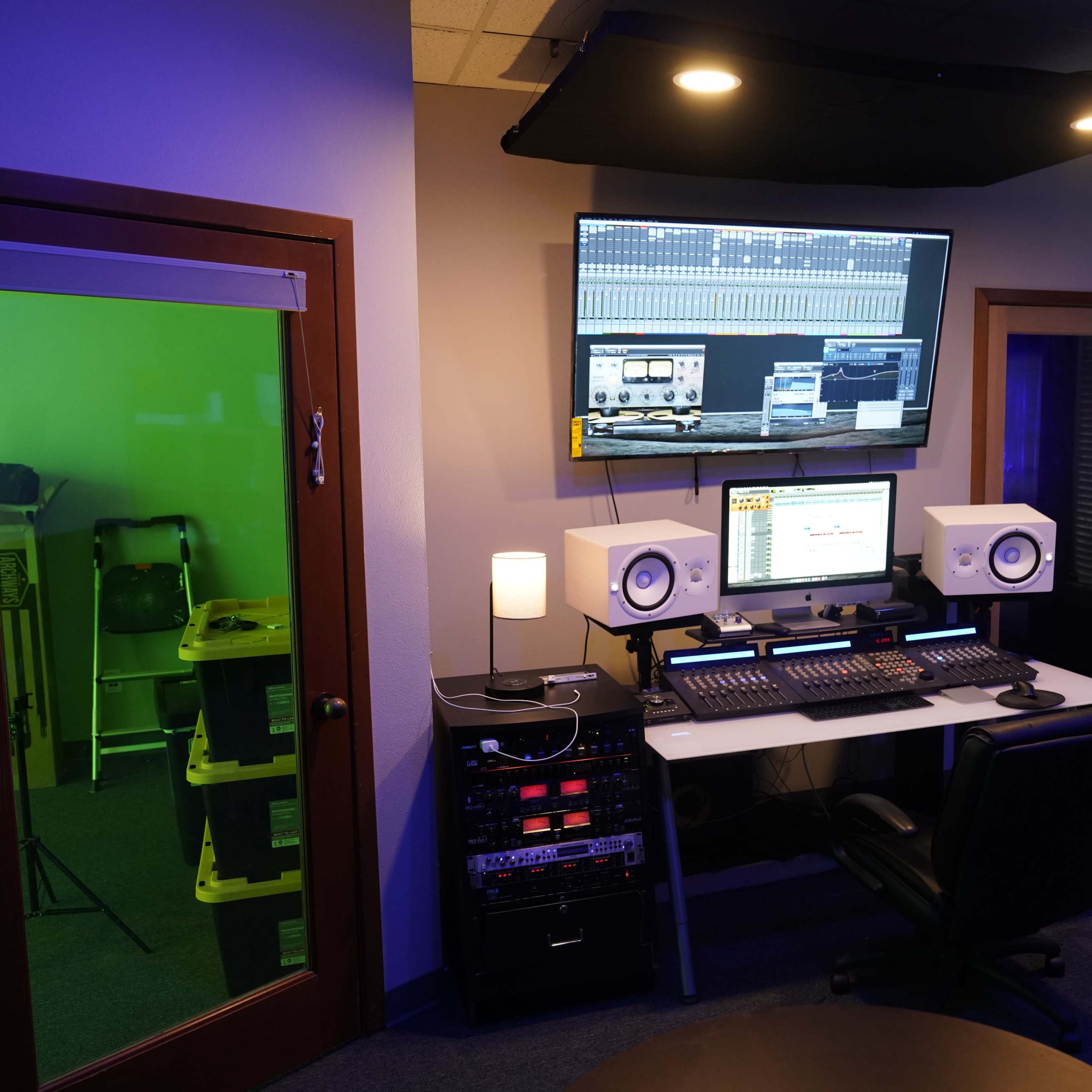 A modern recording studio features a desk with audio equipment, two monitors, and a green room visible through a glass door.