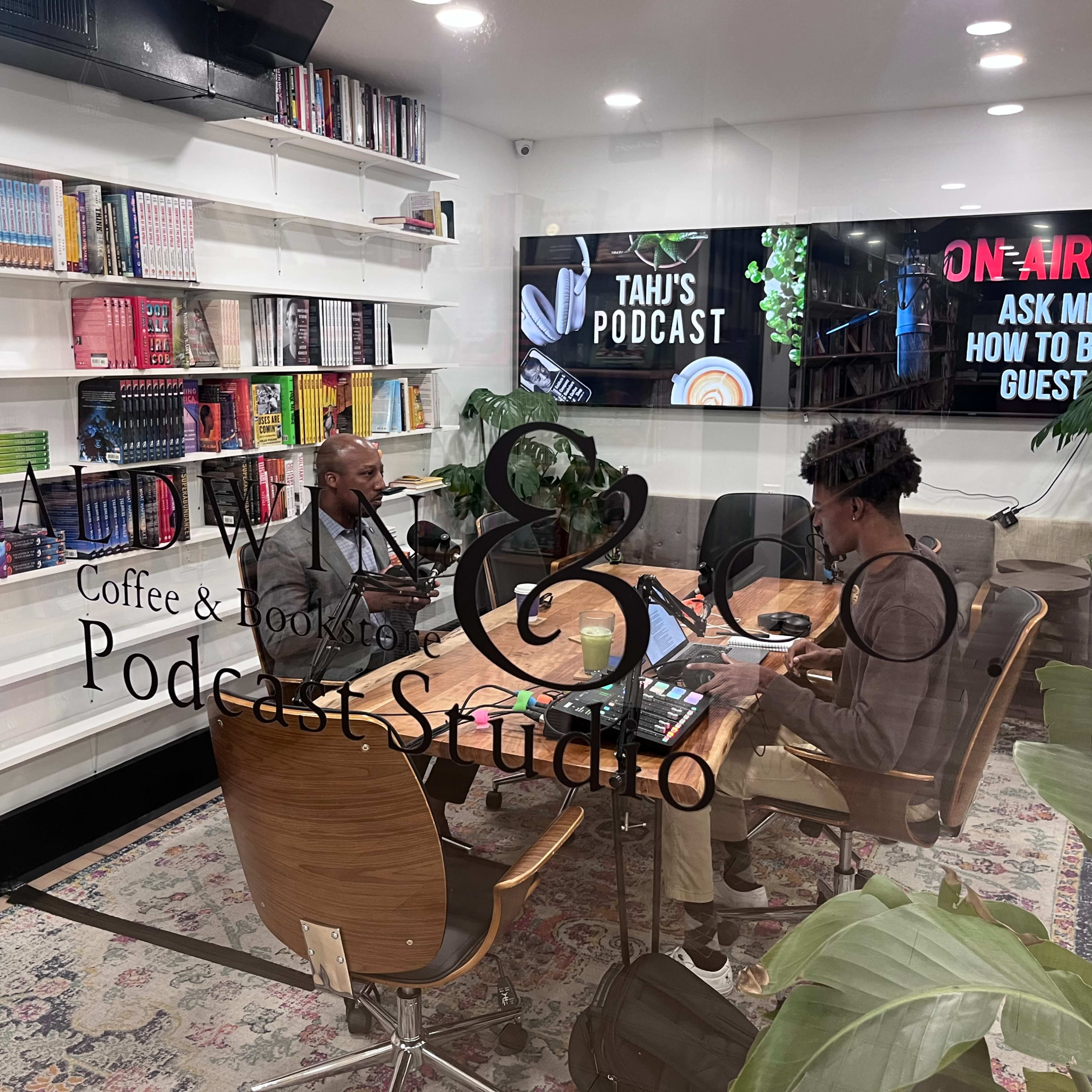 A man and a woman sit at a table in a podcast studio surrounded by books and a large screen displaying podcast information.