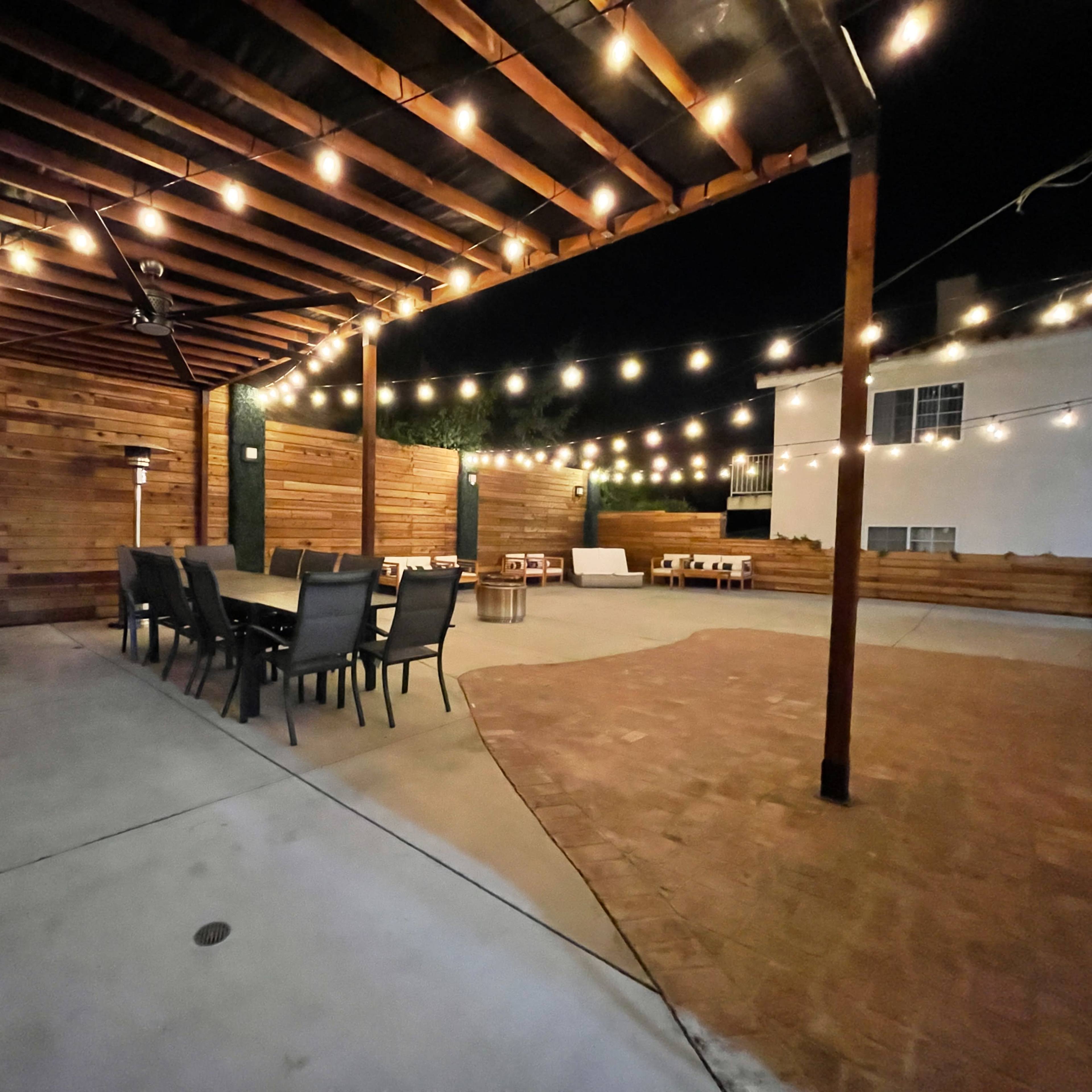 A spacious outdoor patio area with wooden walls, string lights overhead, and seating arrangements for dining and lounging at night.