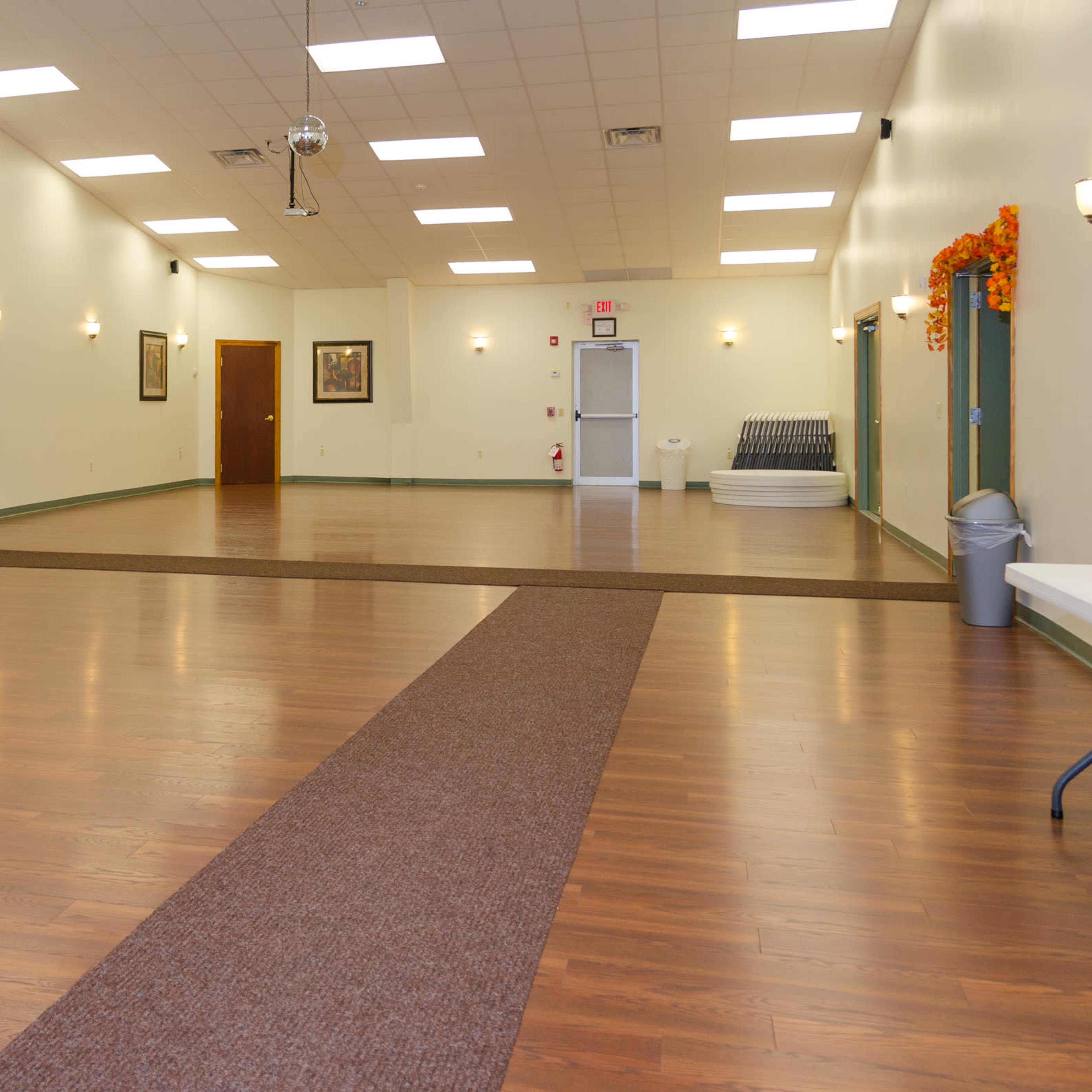 A spacious indoor venue with wooden flooring, green walls, and minimal furnishings, featuring a central carpeted walkway.