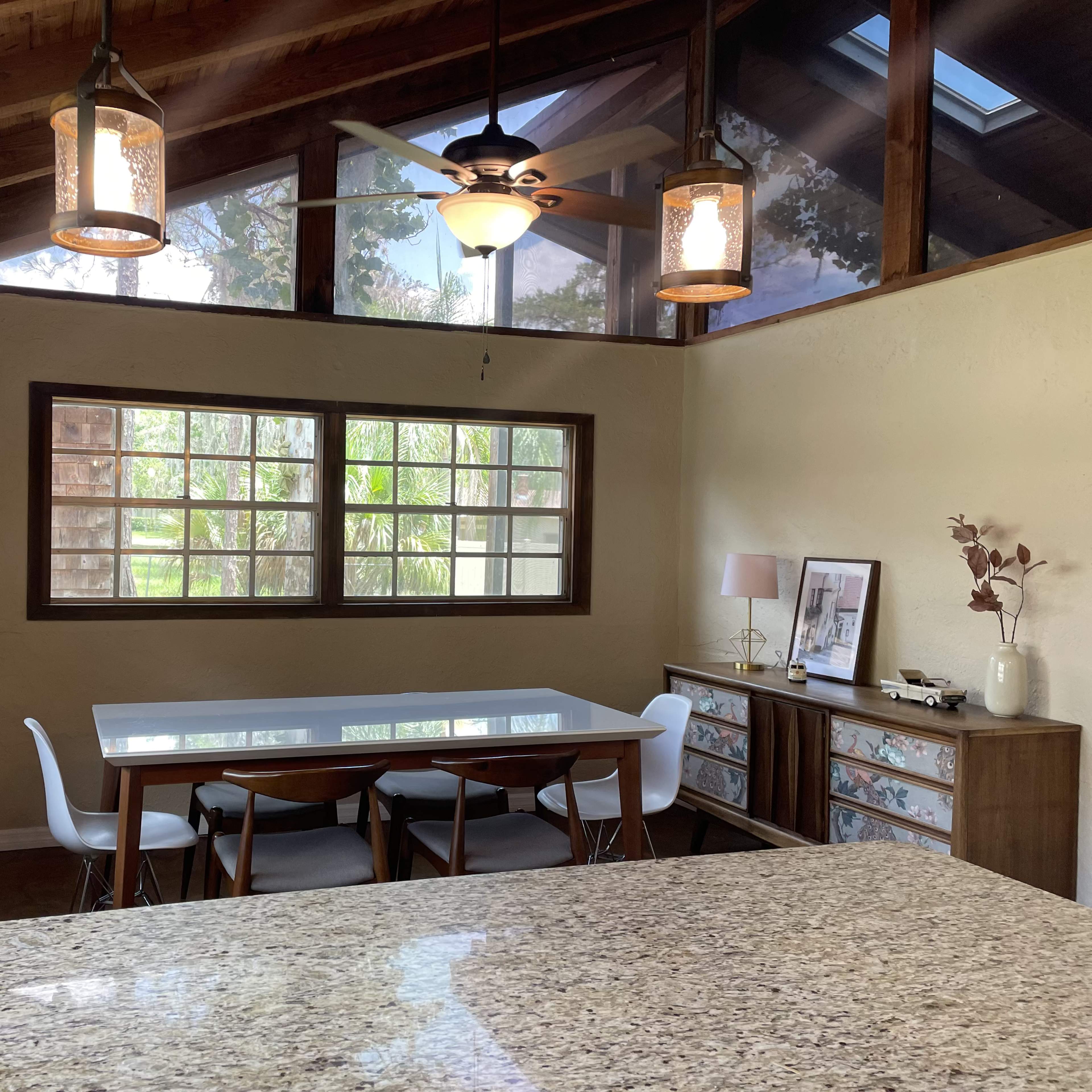 A dining area with a rectangular table surrounded by chairs, situated under a vaulted ceiling with large windows allowing natural light to enter.